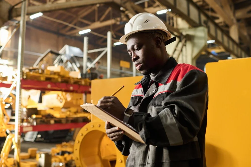 A man wearing a white safety helmet and a black and red work jacket writing on a clipboard inside an industrial warehouse with yellow machinery and metal shelving in the background.