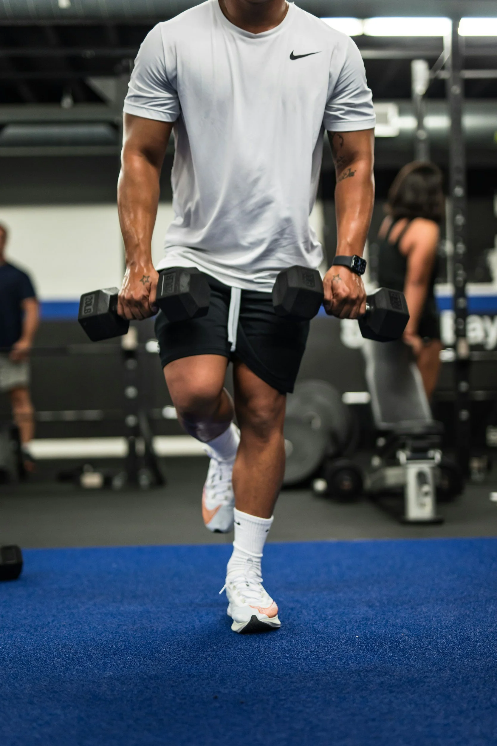A man in a white Nike shirt and black shorts works out with dumbbells in a gym, lifting his right knee while balancing on his left leg.