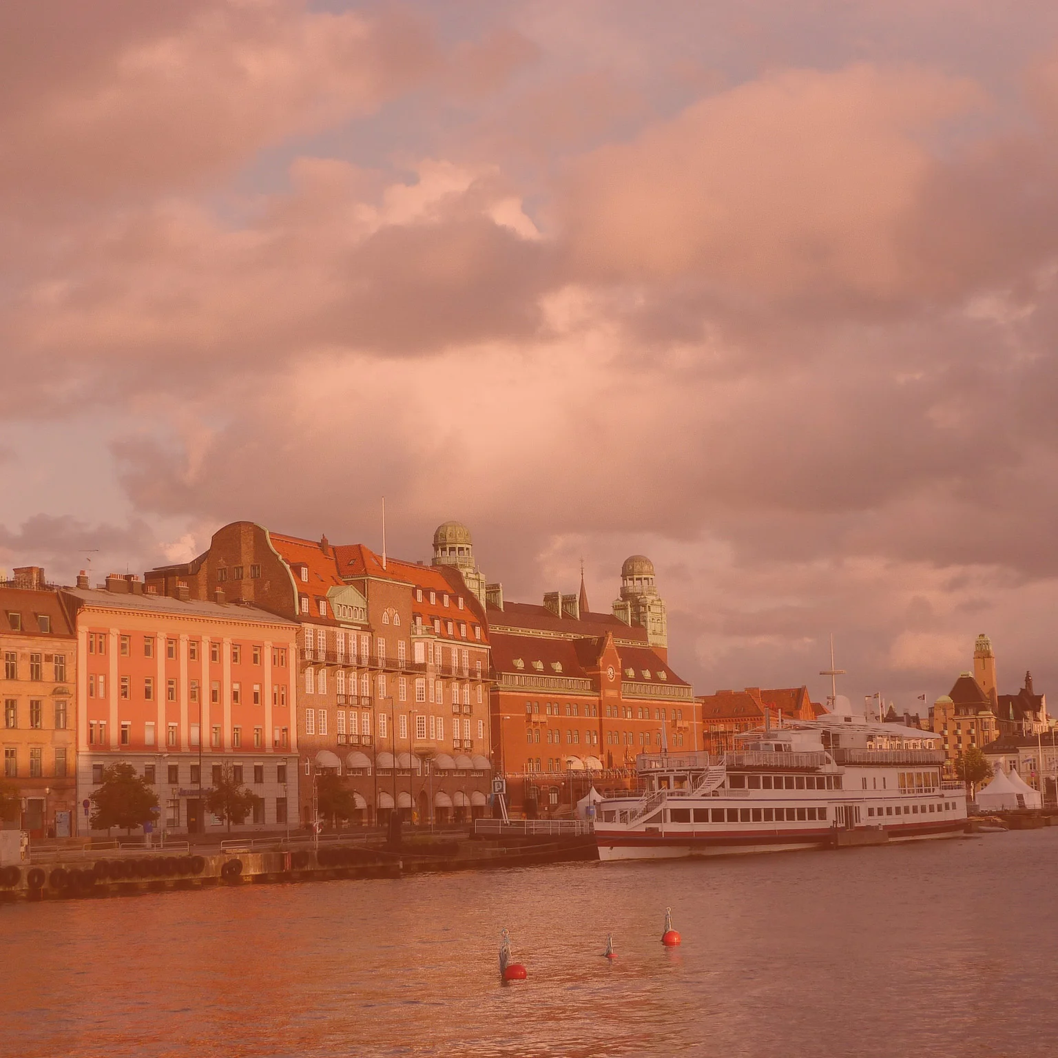Photo of historic waterfront buildings and a boat docked along the river, under a sky with scattered clouds, taken during sunset.