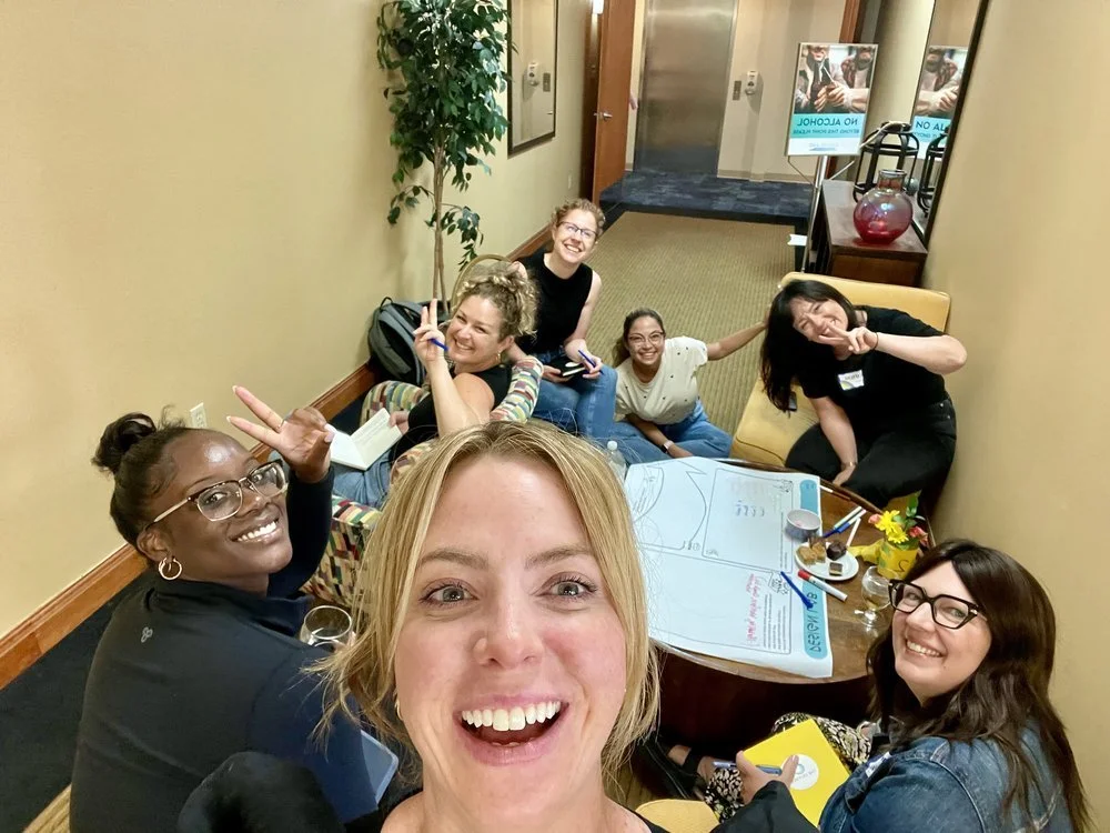 A group of seven women gathered around a table in a hallway, smiling and making peace signs, with a whiteboard, markers, and snacks on the table.
