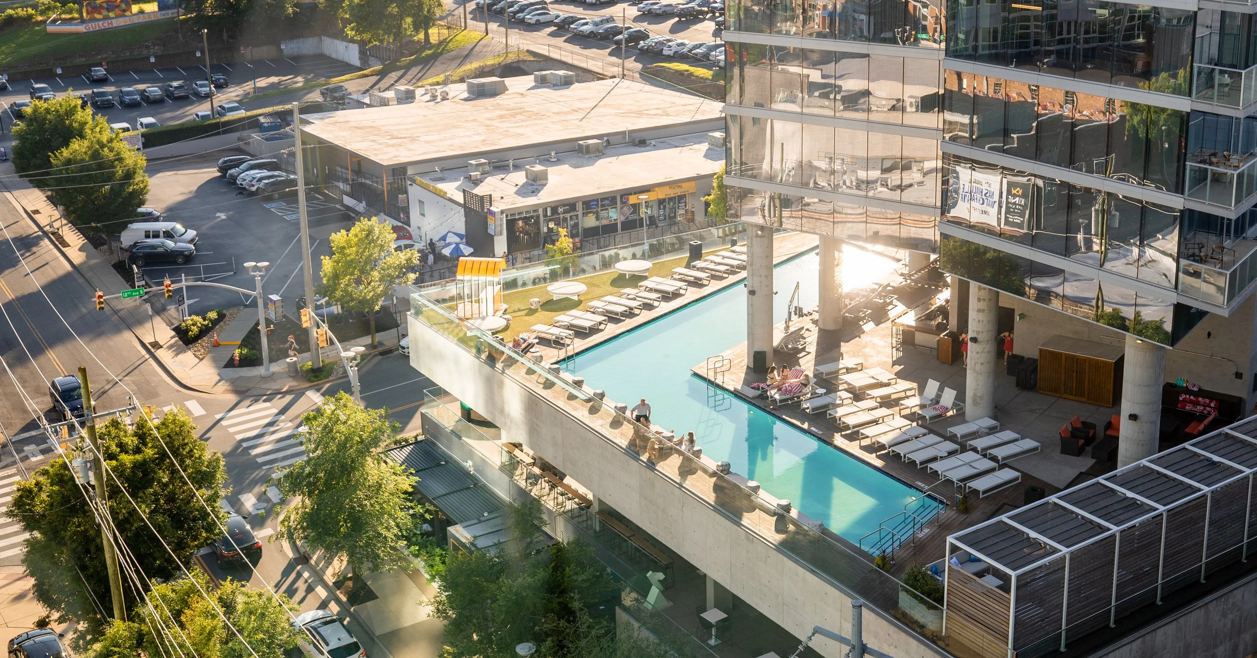 Aerial view of a modern building with a rooftop pool and lounge area. Surrounding streets are busy with cars and traffic lights. There is a parking lot and trees nearby.