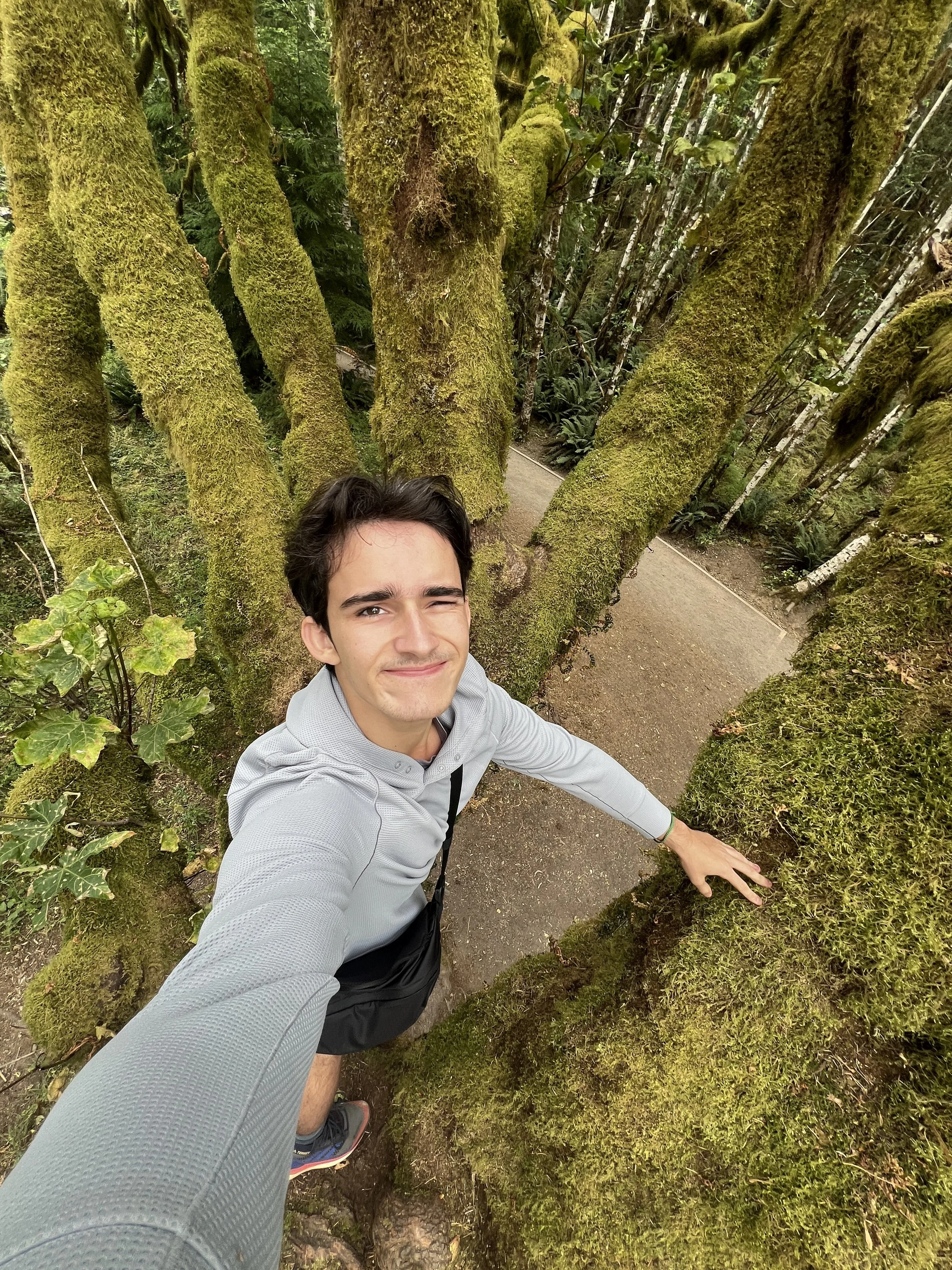 Young man taking a selfie with a wide-angle lens on a trail surrounded by moss-covered trees in a lush green forest.