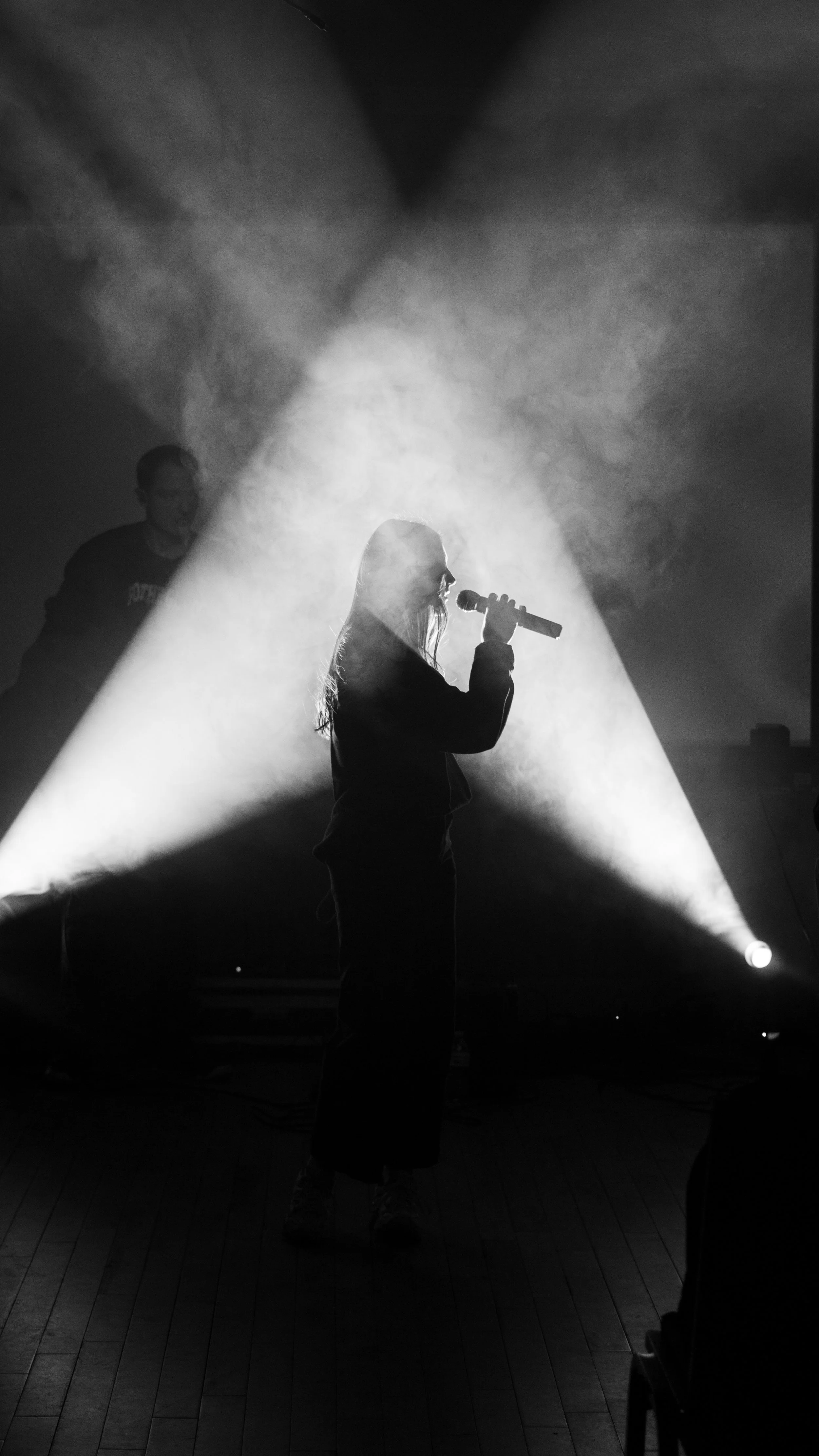 A black and white photo of a woman singing into a microphone on a stage, with dramatic lighting and a person behind her.