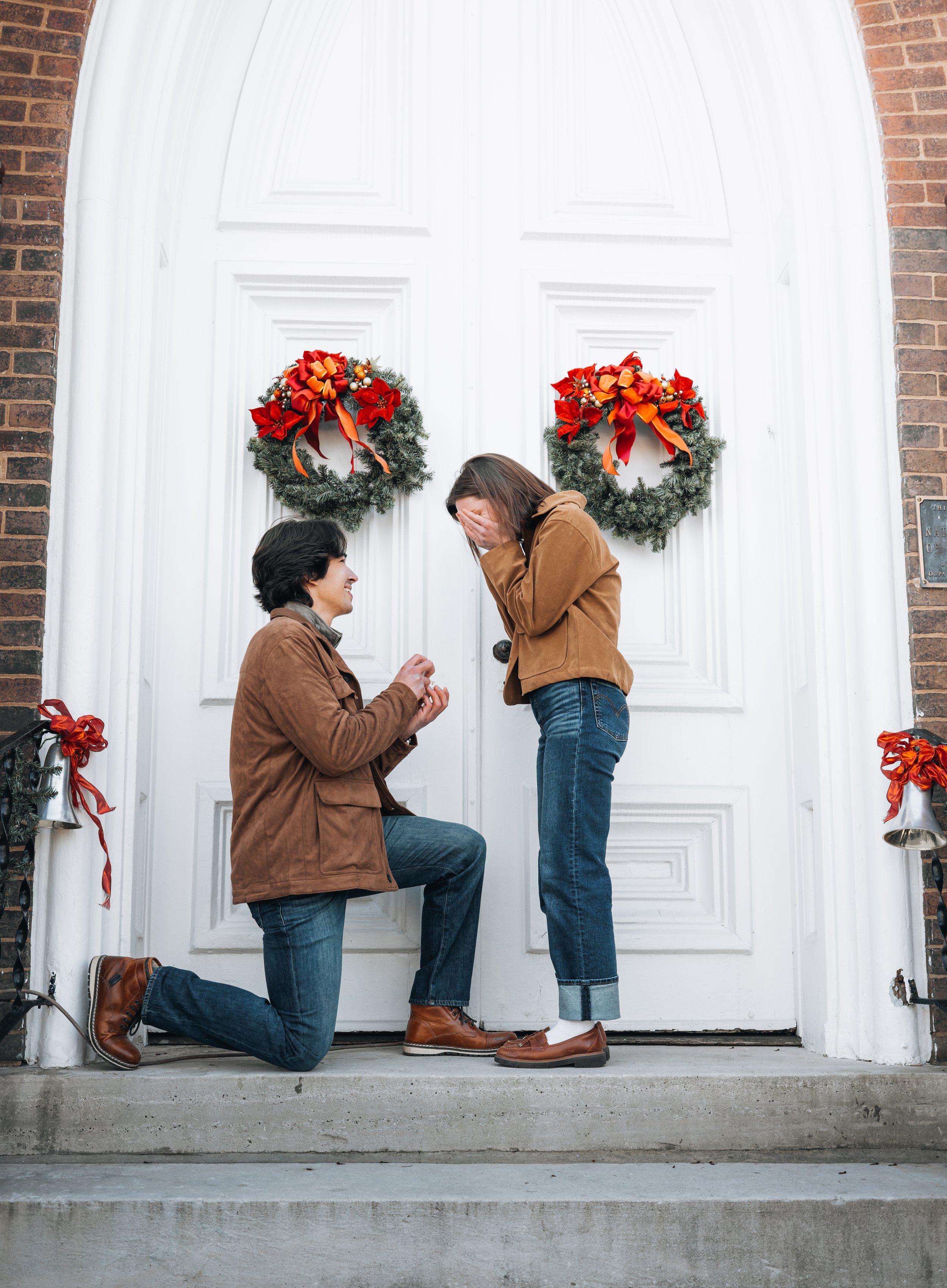 A man is proposing marriage to a woman in front of a decorated door with wreaths and bells, while she reacts happily with her hands covering her face.