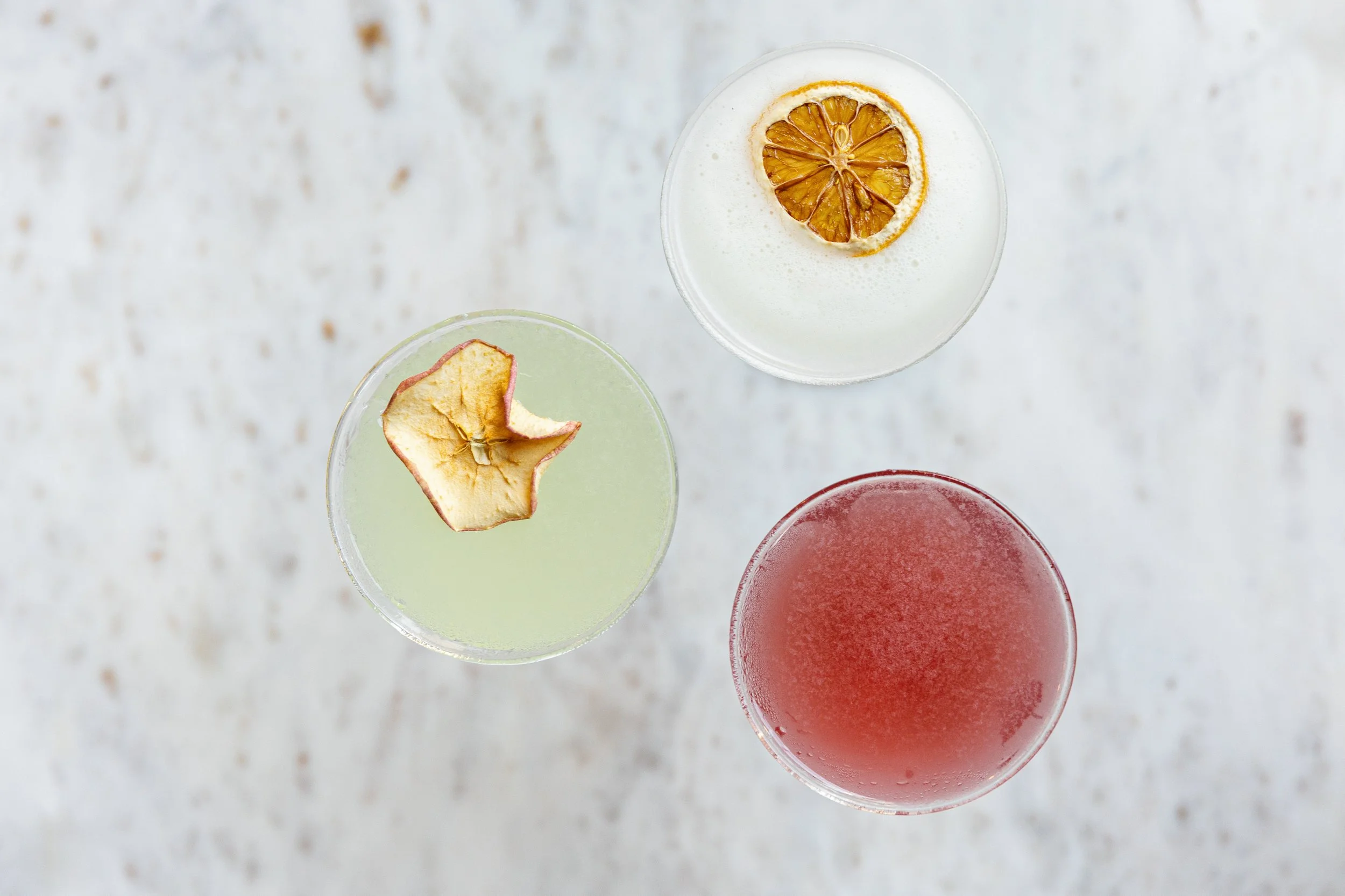 Top-down view of three cocktails with dried fruit garnish on a white textured surface.