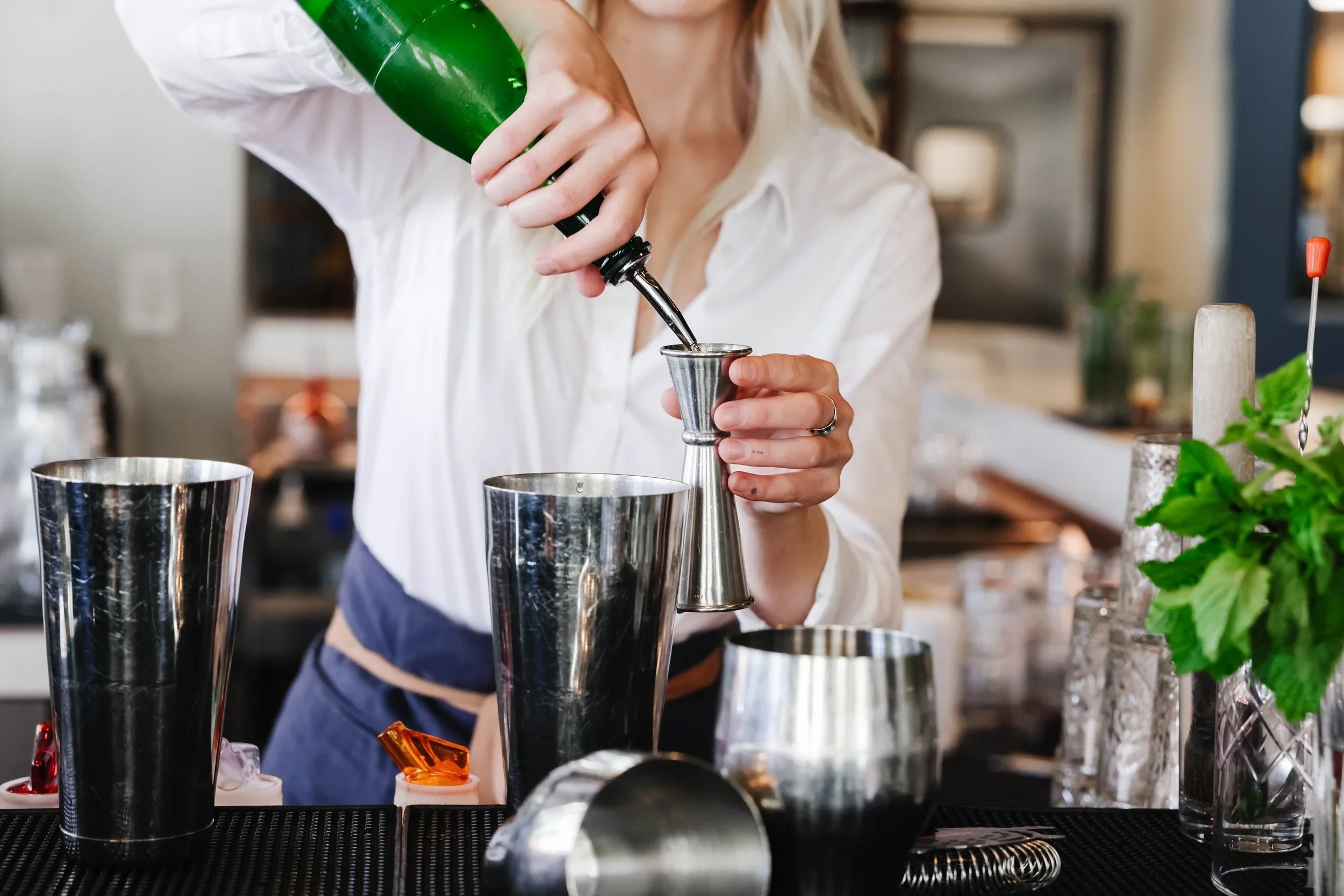 A bartender pouring a dark liquid from a green bottle into a metal jigger at a bar counter with various bar tools and plants in the background.