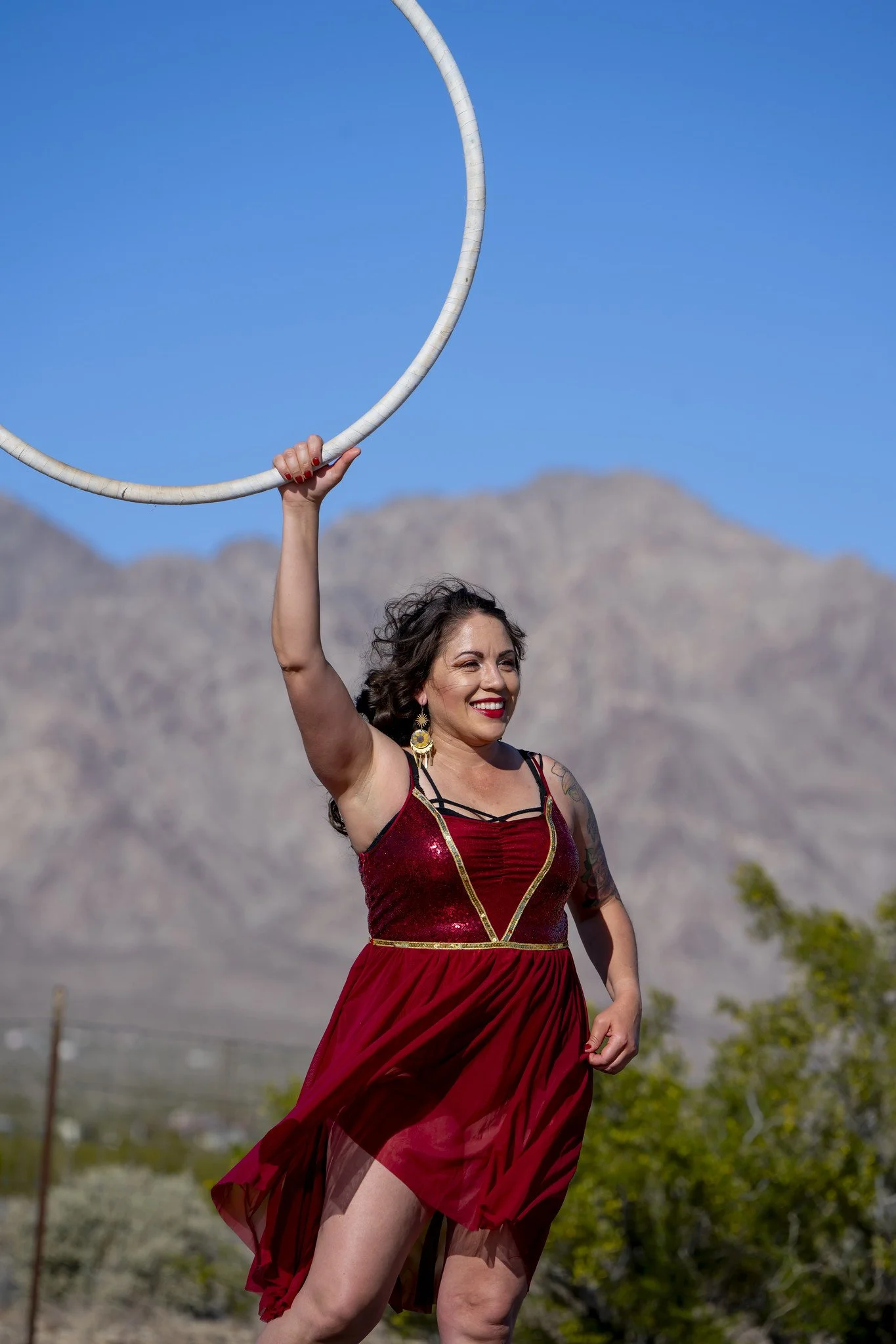 A woman dressed in a red dress with gold accents is holding a large hoop above her head outdoors. She has dark hair and is smiling, with a mountainous landscape and blue sky in the background.