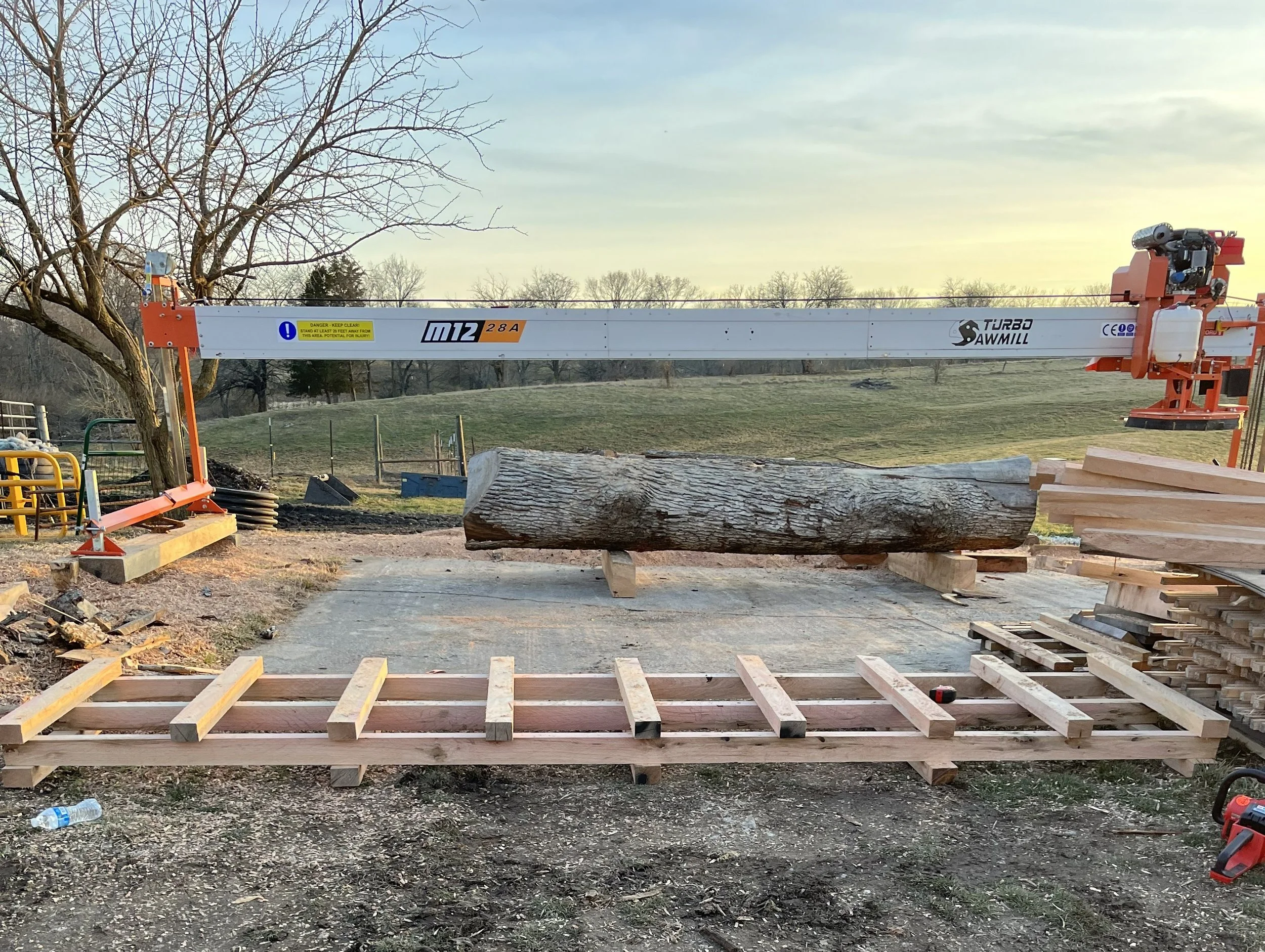 A large log resting on wooden supports with a wood cutting sawmill in the background, set outdoors on a dirt and gravel surface with trees and a grassy field under a cloudy sky.