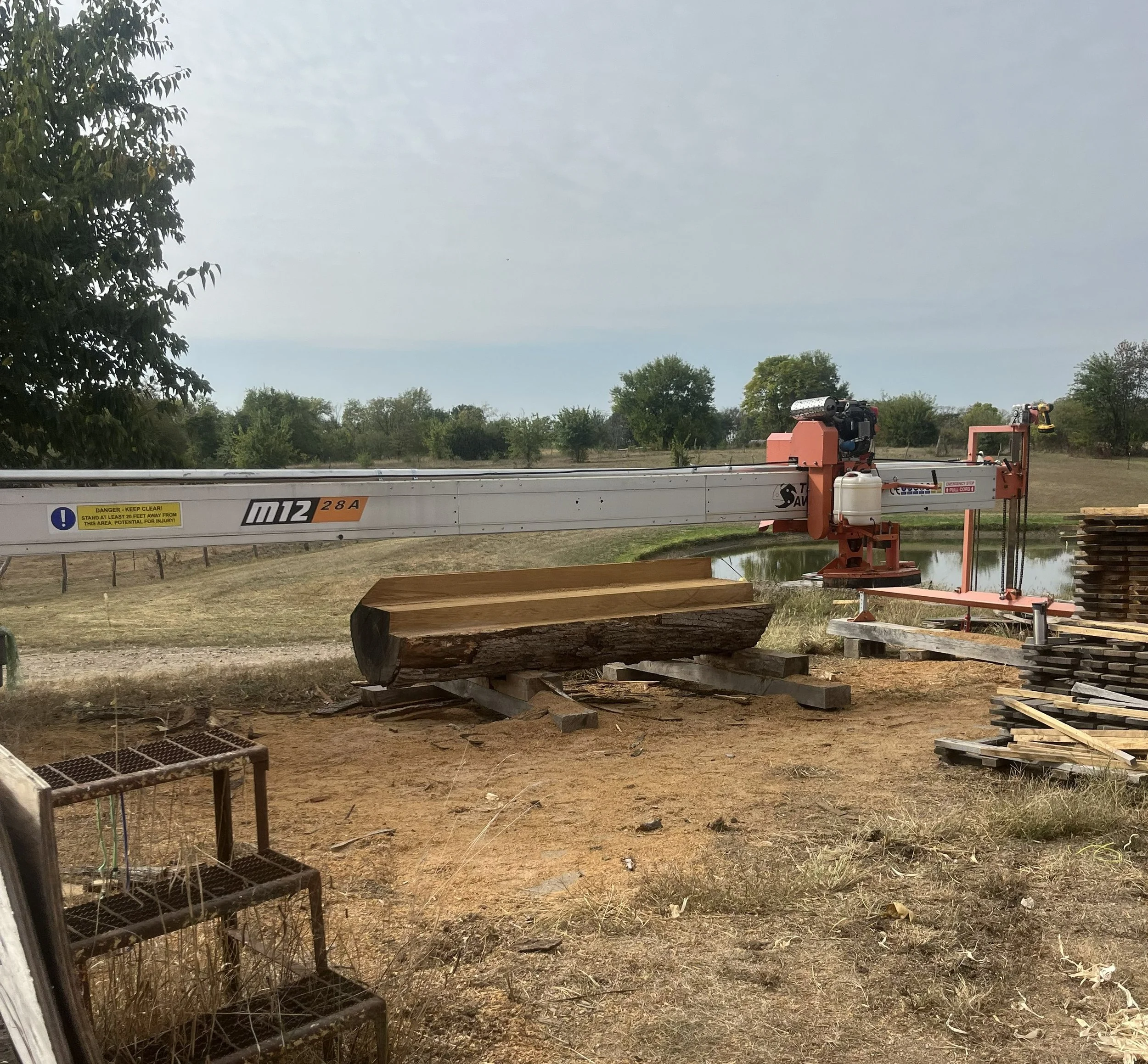 A large orange and white hydraulic sawmill cutting a log into lumber at a woodworking yard near a pond with trees in the background.