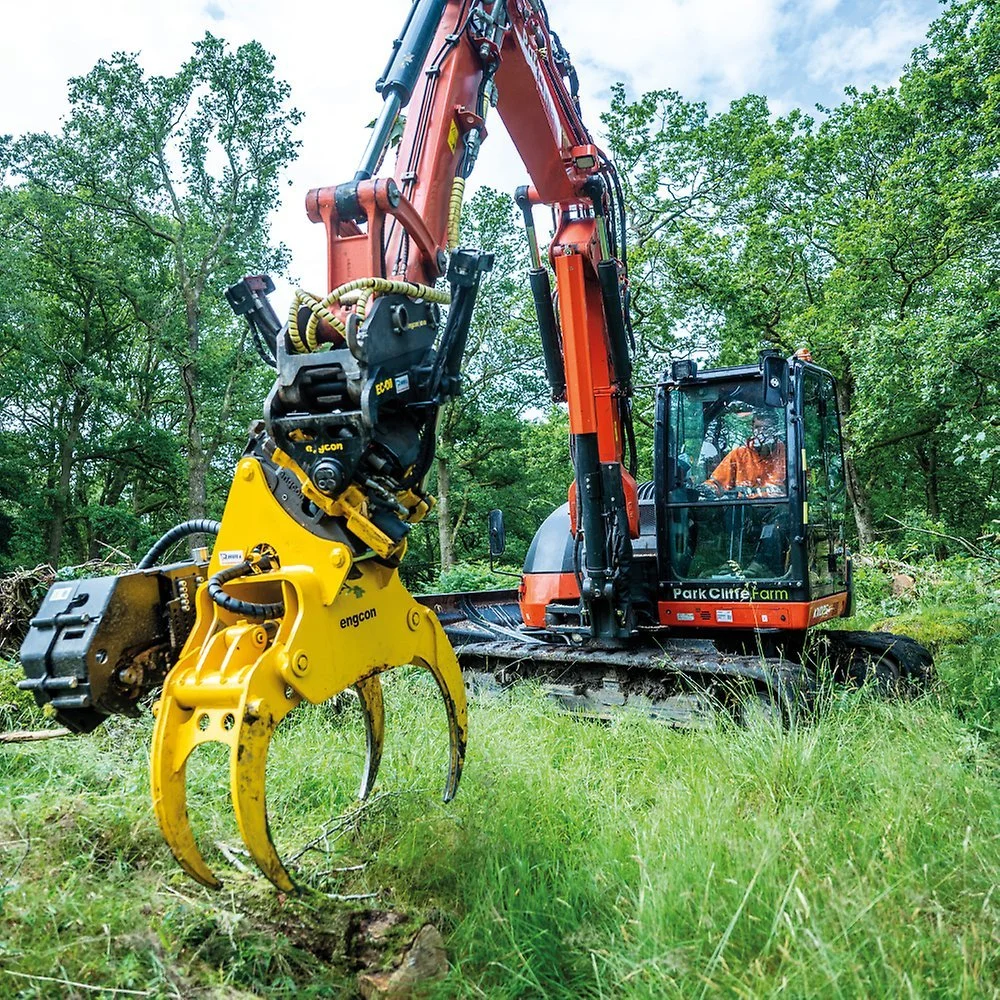A large orange and black excavator with a yellow grapple attachment working in a green wooded area.