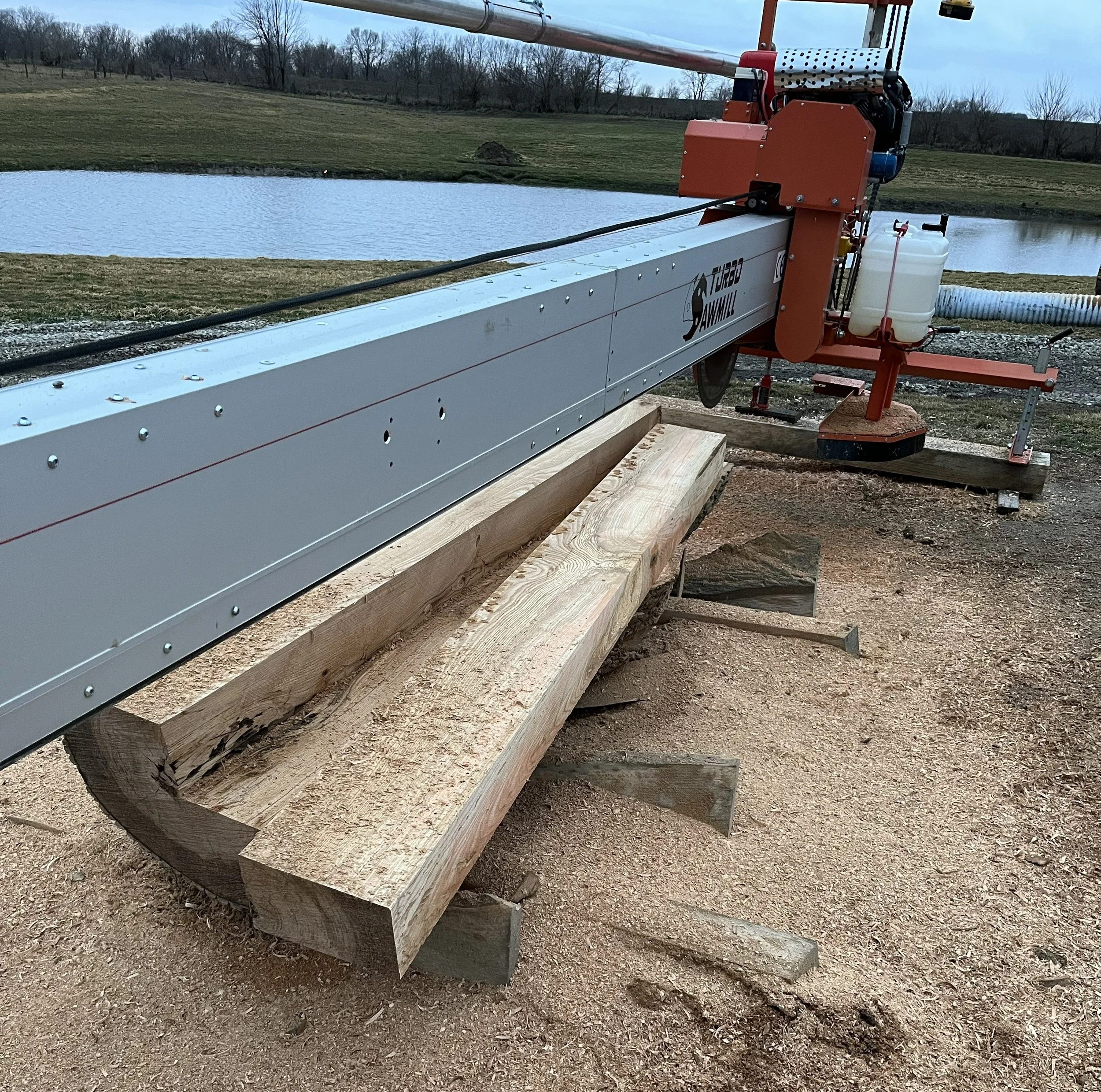 A large sawmill machine with a long metal arm cutting a thick wooden beam outdoors near a pond, with trees and grassy fields in the background.