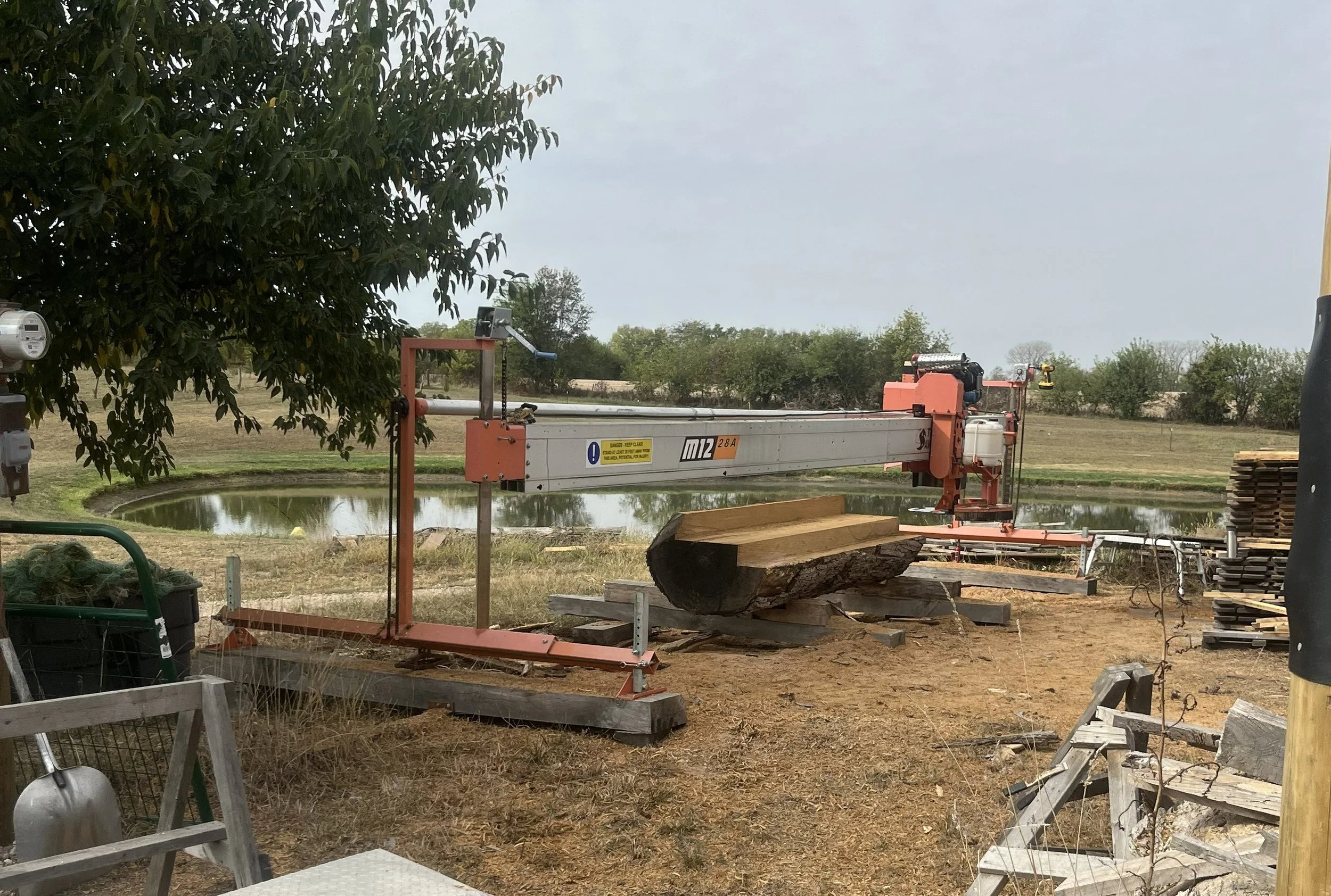 A large log with freshly cut top standing on supports, next to a wood cutting mill on a grassy yard with a pond and trees in the background.