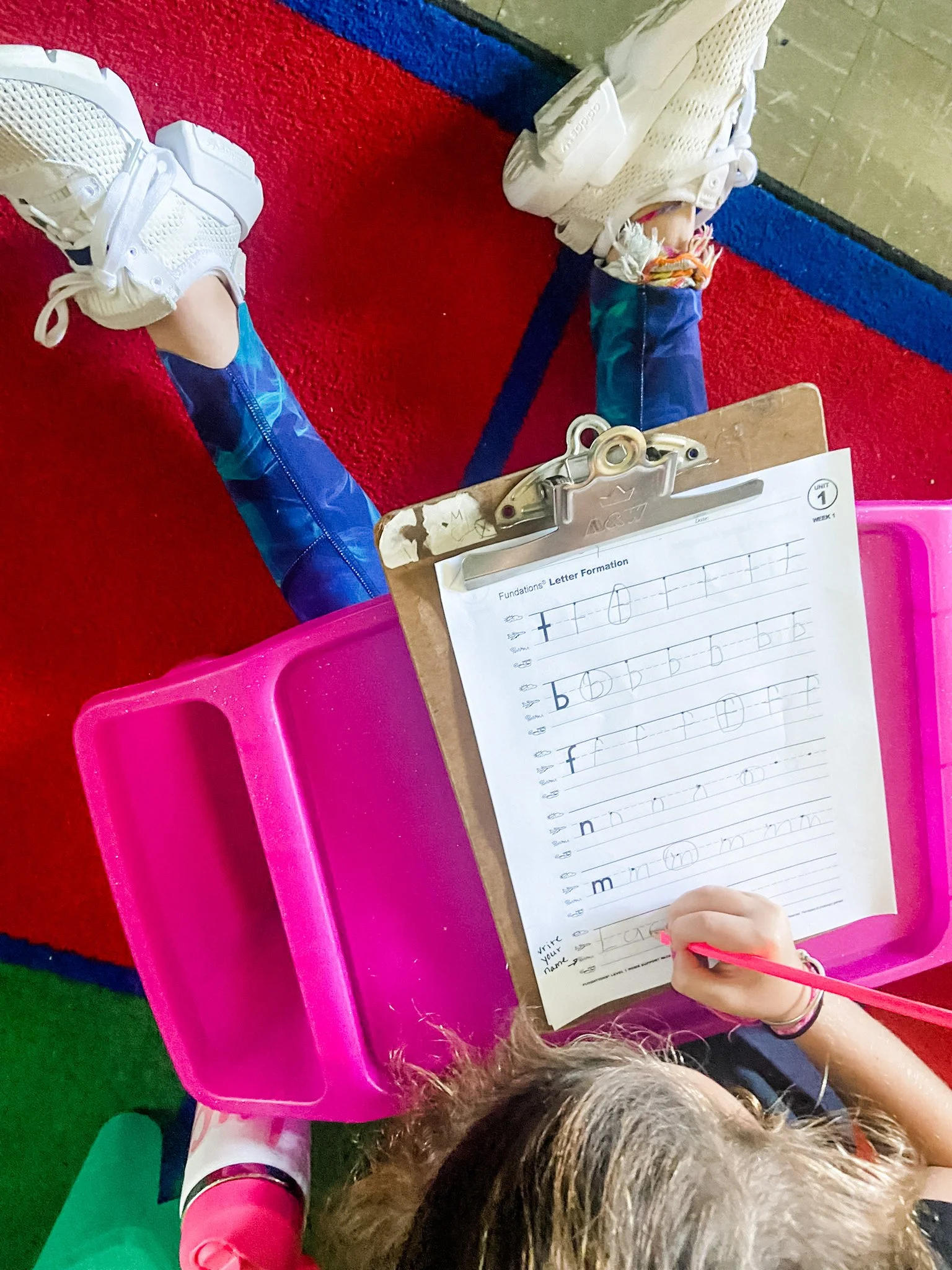 Child seated at a pink desk practicing cursive letter formation on a worksheet, wearing white sneakers with colorful socks, with a clipboard and colorful carpet in the background.