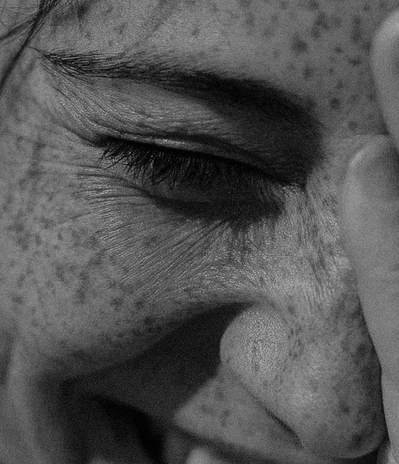 Close-up black and white photograph of a person's face with eyes closed, focusing on the area around the eye, eyelid, eyelashes, and the skin texture.