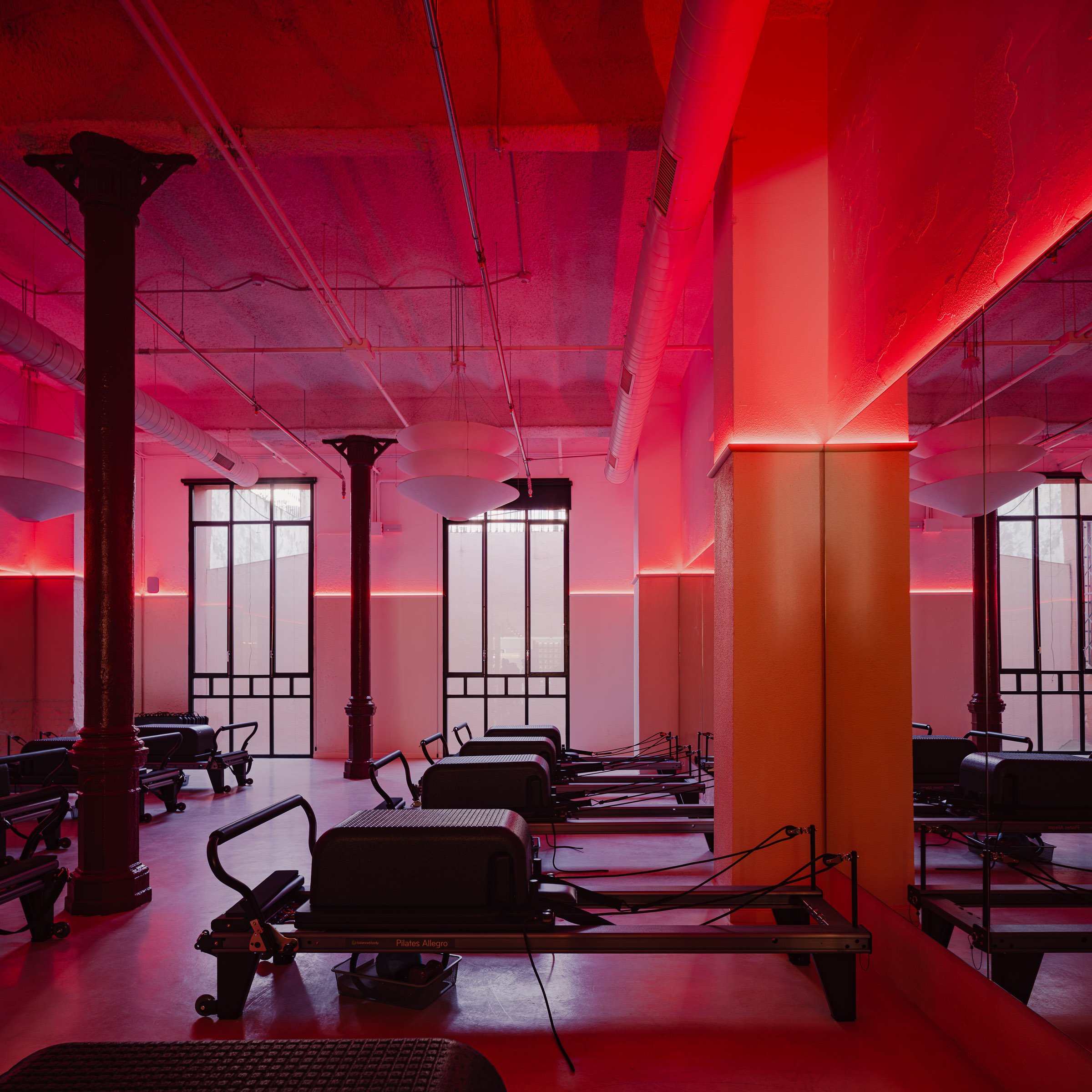 Pilates reformer machines in a fitness studio with pink and red lighting and large windows.