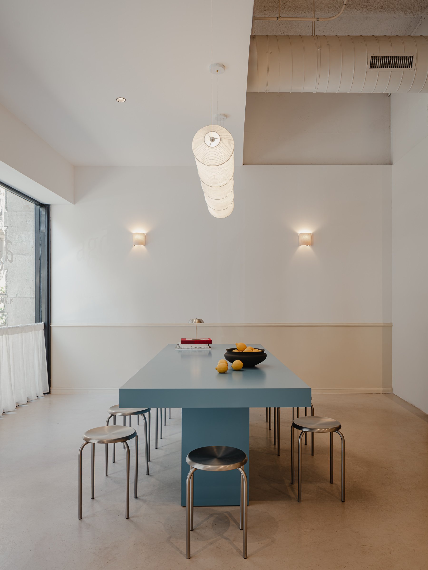 Minimalist dining area with a blue rectangular table, six metallic stools, black bowls with lemons, and a scattered lemon on the table. White walls with two small wall-mounted lights, a large window, and a modern ceiling light fixture.