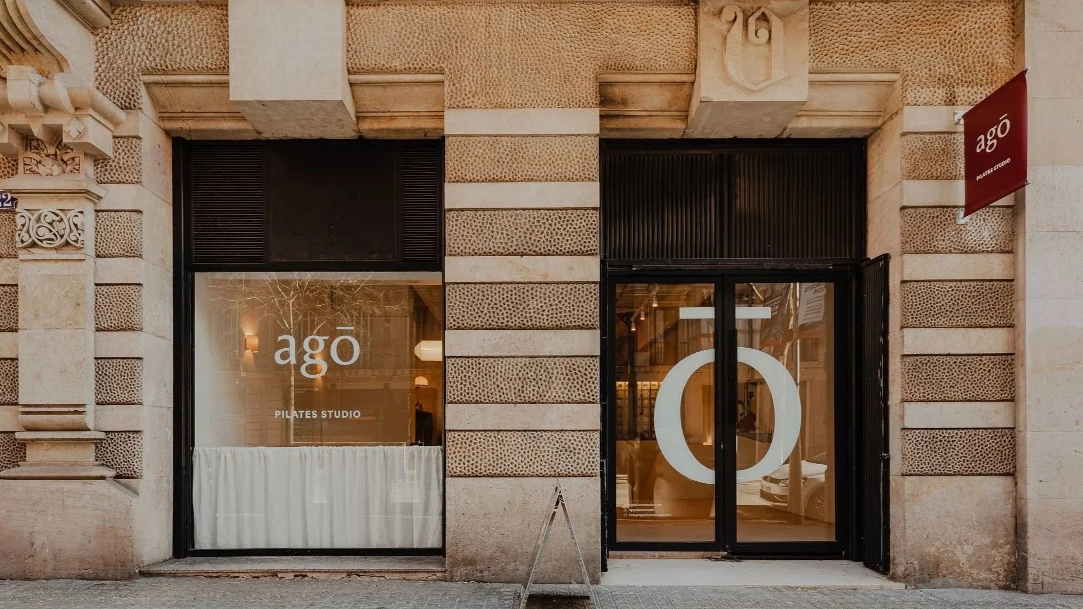 Exterior of a Pilates studio named 'Ago' with a large window and glass door, showing the interior, and a small signboard outside on a city street.
