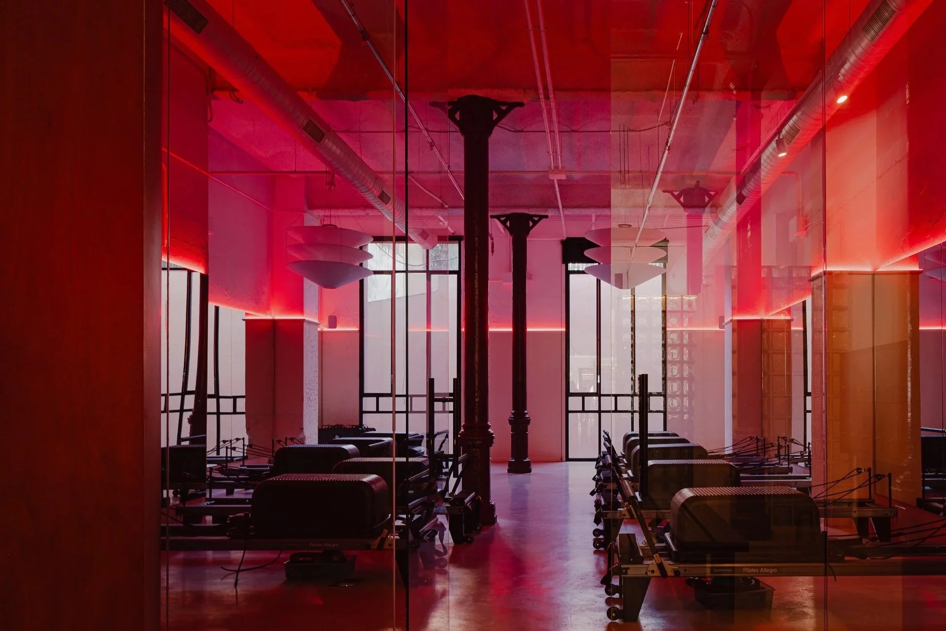 Reformer pilates space with reformer beds, mirrors, and ceiling lights, illuminated with red and pink lighting.