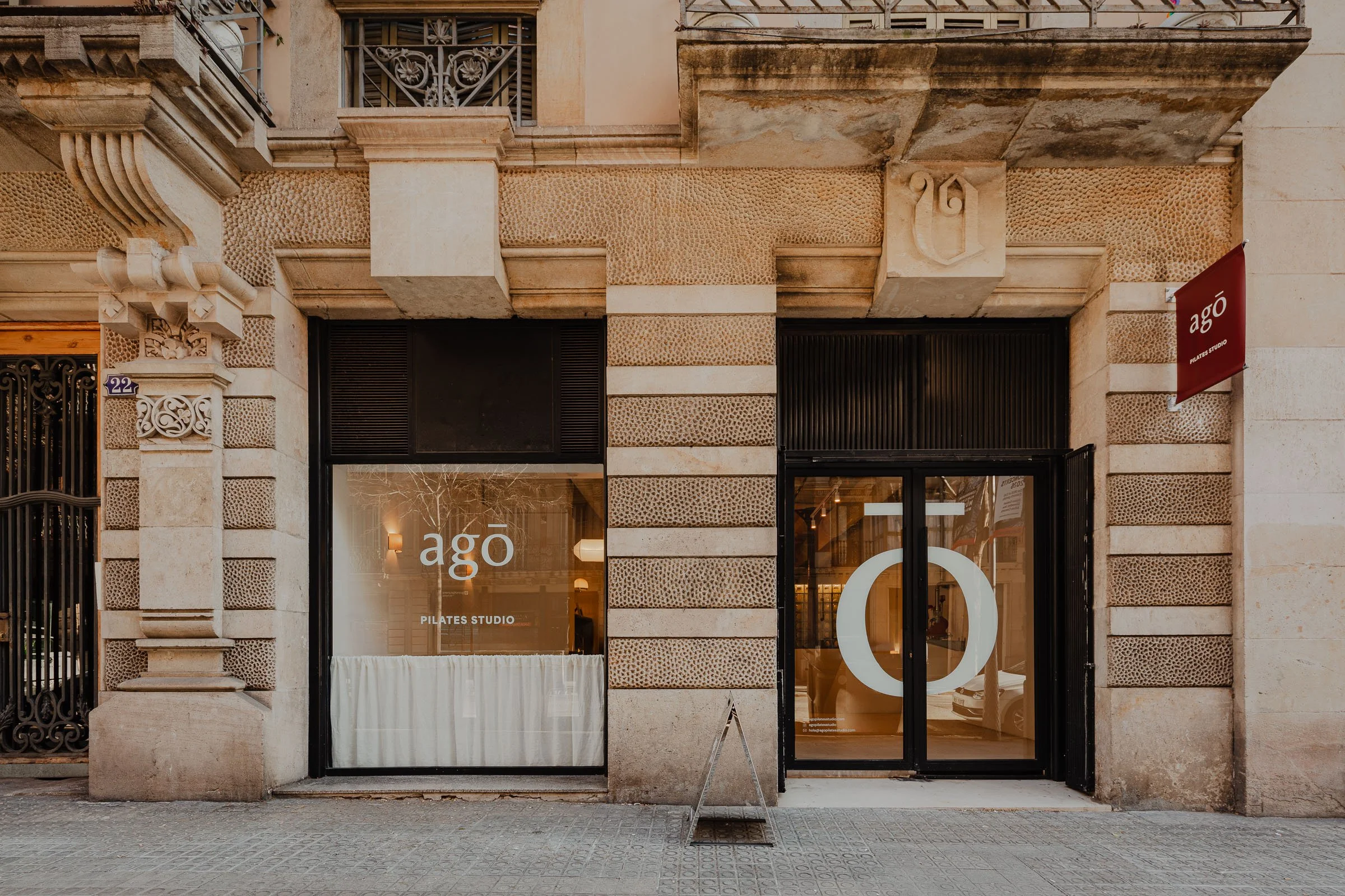 The storefront has a beige stone facade with textured patterns, decorative architectural elements, and black frames around large windows and door. A red sign to the right reads 'agó' with smaller text underneath. Through the windows, indoor lighting and a white curtain are visible.