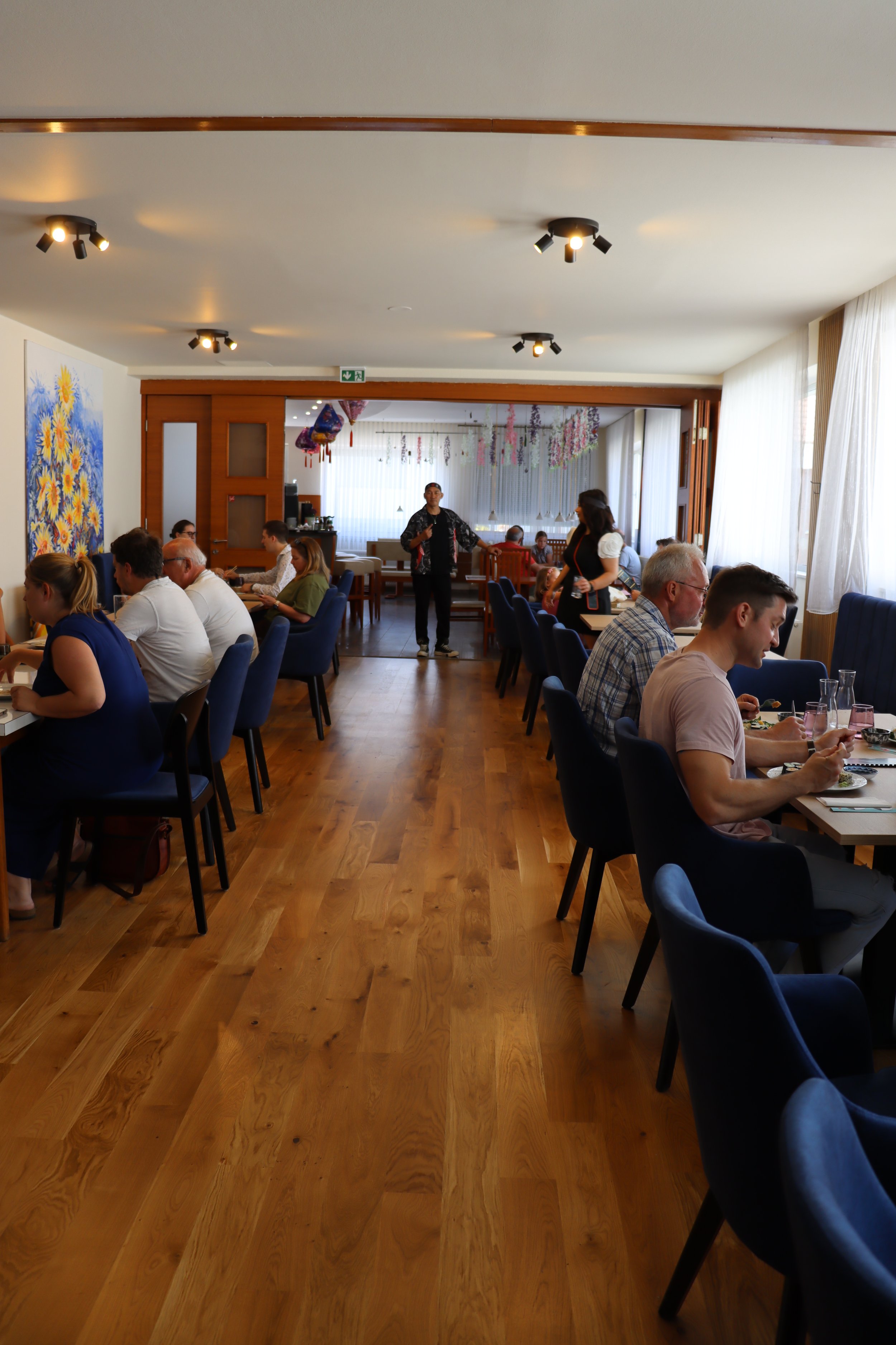 Innenaufnahme eines Restaurants oder Cafés mit mehreren Gästen, die am Tisch speisen, und einer freundlichen jungen Frau, die im Hintergrund einen Wasserkrug hält, während ein Mann in einem schwarzen Hemd und einer schwarzen Jacke spricht.