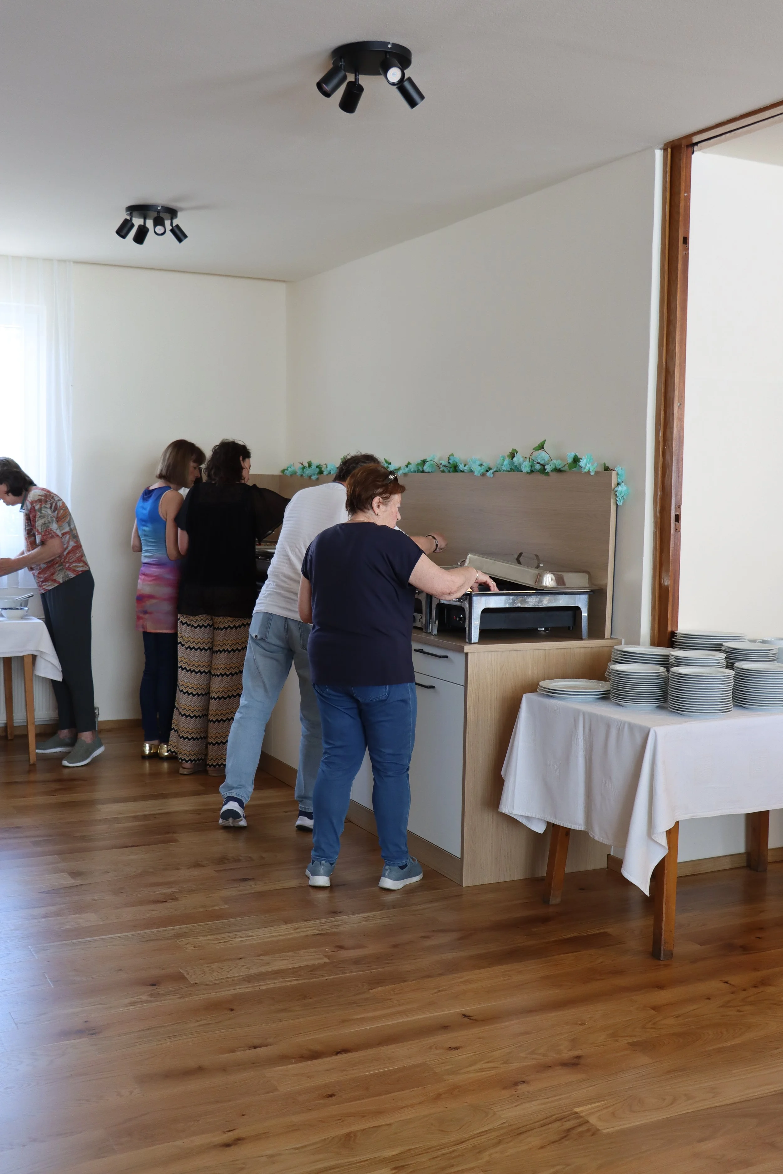 Menschen stehen in einer Reihe an einem Buffet, um sich Essen zu holen, im Hintergrund steht ein Buffet mit Tellern, es ist eine Gesellschafts- oder Familienfeier in einem Raum mit Holzboden.