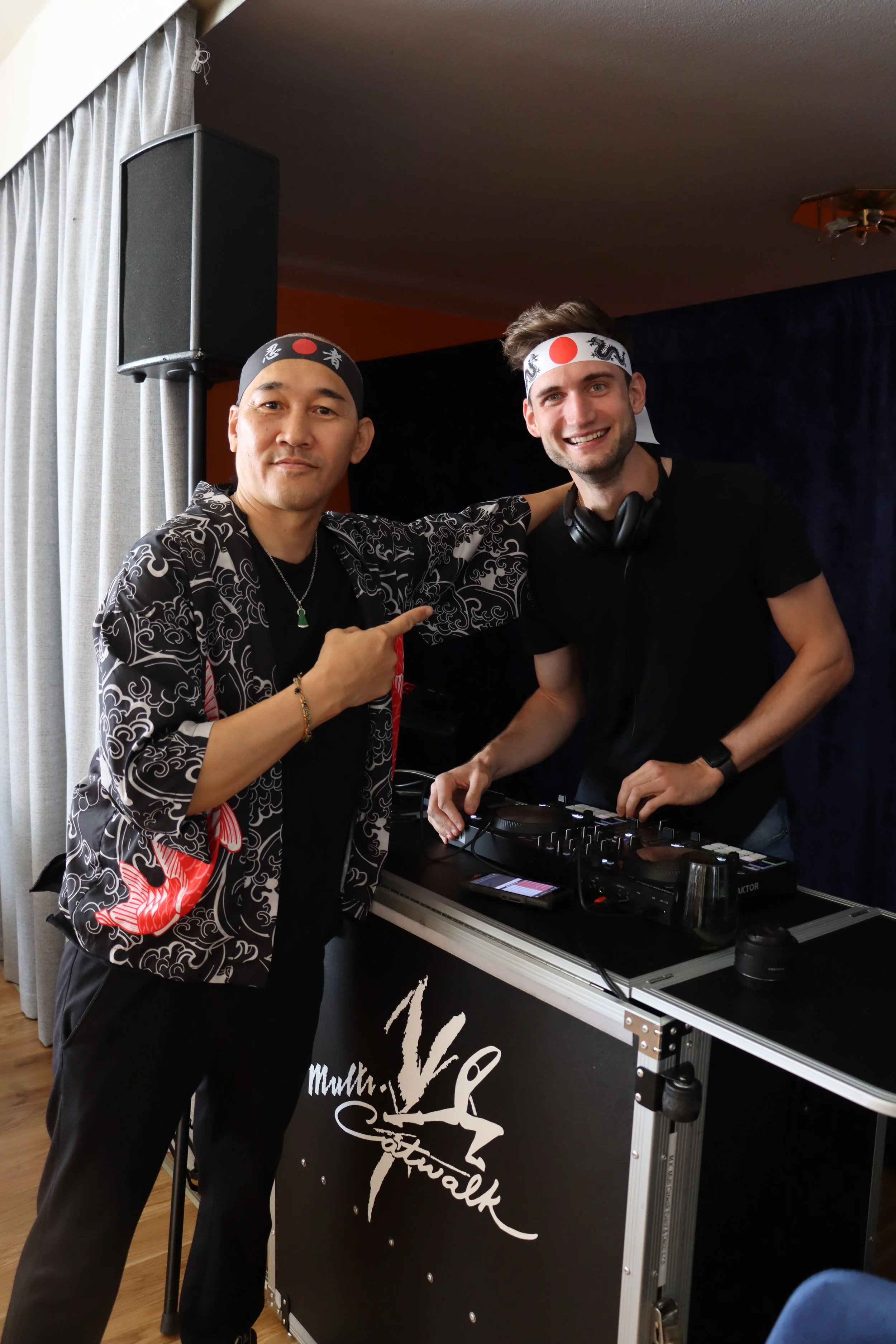 Zwei Männer, die eine Party oder ein Event feiern, tragen japanische Kopfbänder (Hachimaki), sprechen in die Kamera, eines der Männer zeigt auf den anderen, während sie vor einem DJ-Setup stehen.