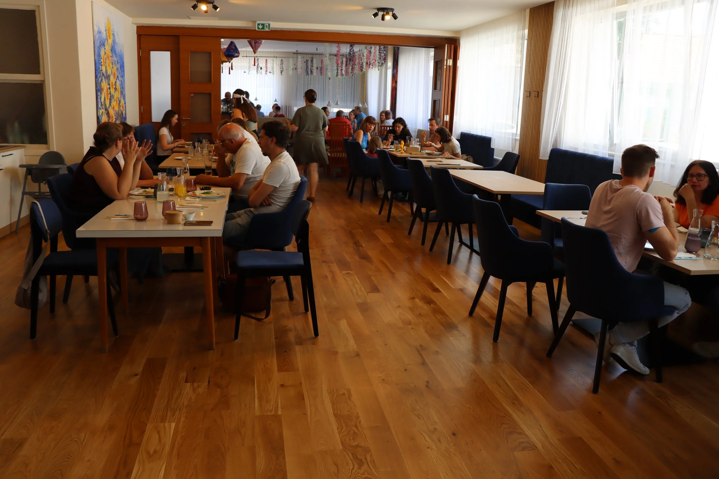 Ein hell erleuchteter Restaurantraum mit mehreren Gästen, die am Tisch essen und sprechen, und Kellnerinnen, die Bestellungen aufnehmen. Die Atmosphäre ist lebendig und freundlich.