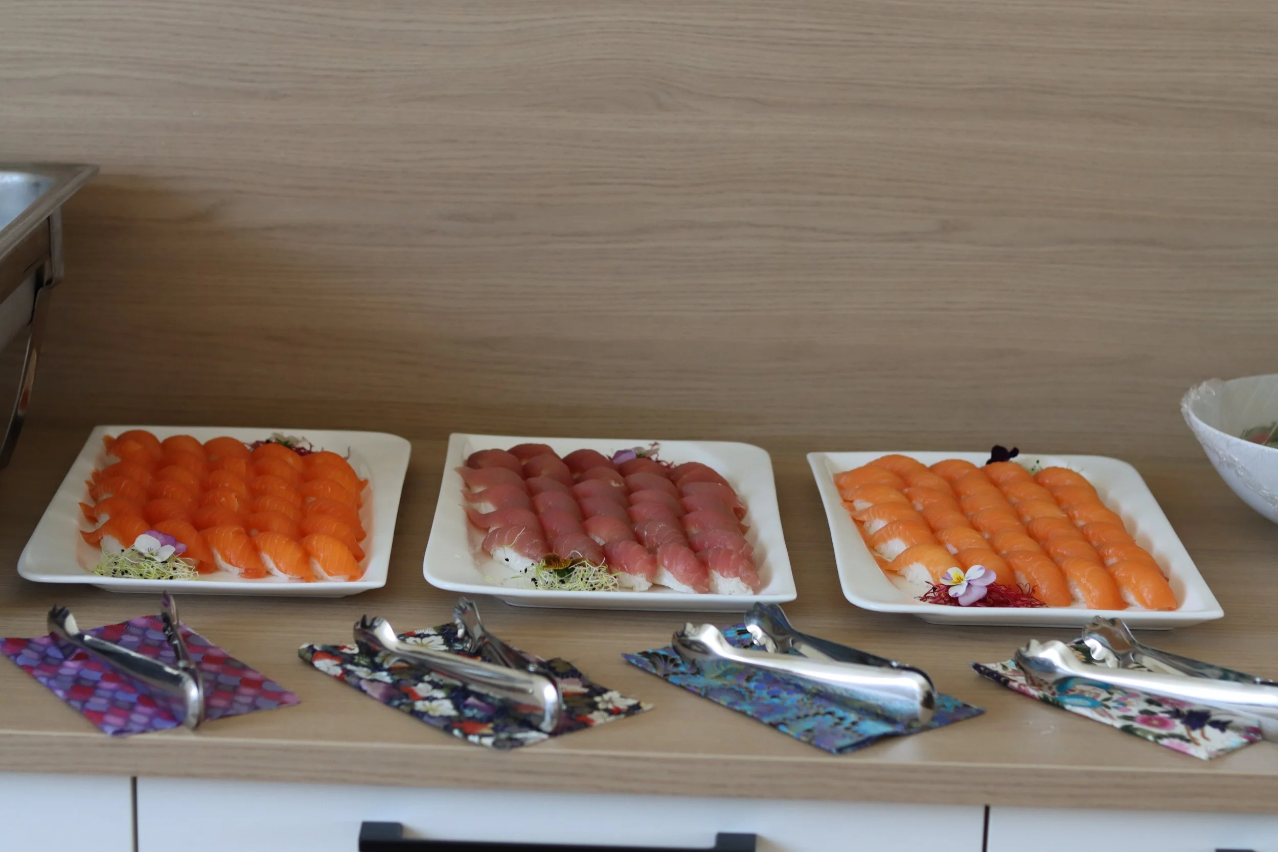 Verschiedene Arten von Sushi auf weißen Tellern, darunter Lachs, Thunfisch und Orange-Lachs-Sushi, mit Dekorationen wie Blumen und Algen, auf einem Holztisch, mit Party-Servietten im Vordergrund.