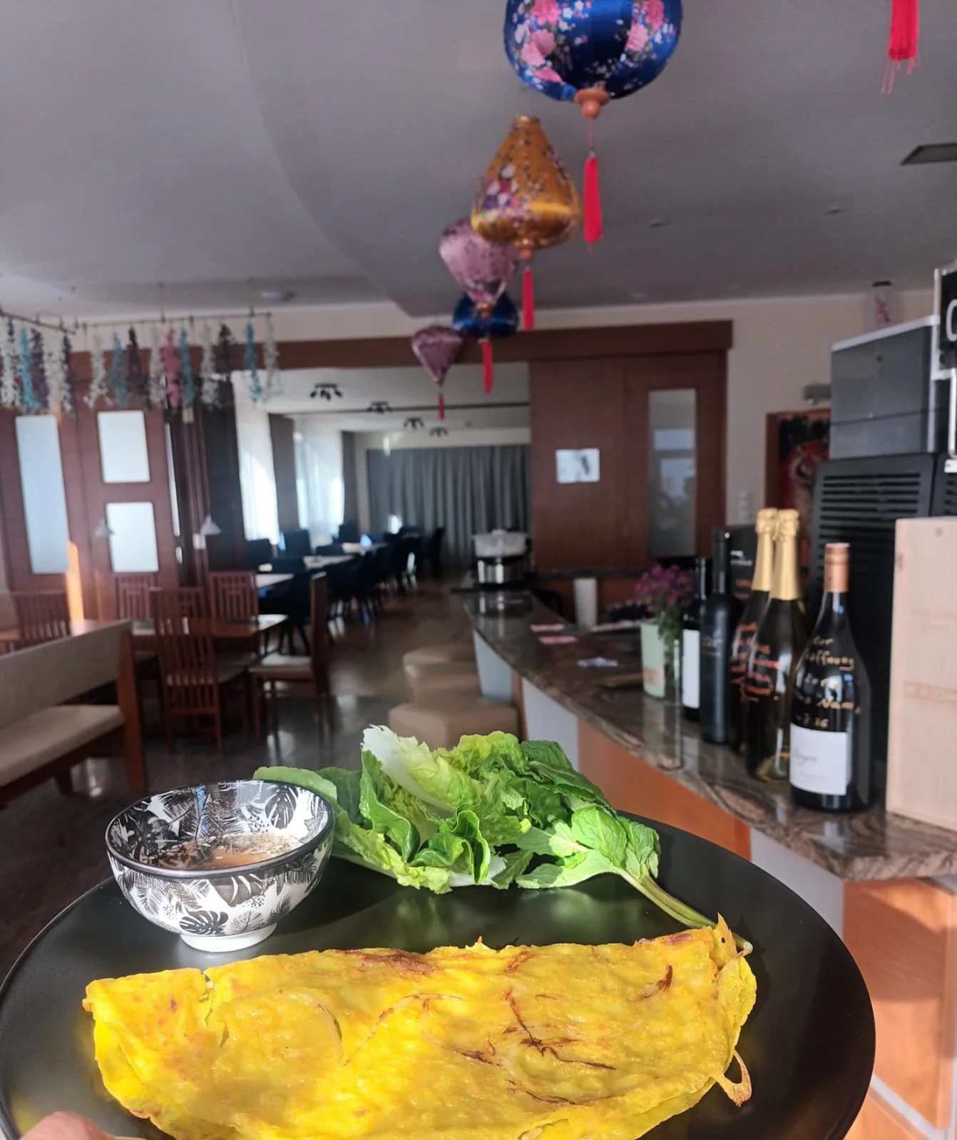 Ein Teller mit einer Omelett-ähnlichen Speise, Salat, einem Glas Wasser und einem kleinen Schälchen mit Sauce auf einem Tisch in einem Restaurant. Im Hintergrund sind Tische, Stühle, Dekorationen, Weinflaschen und eine Bar sichtbar.