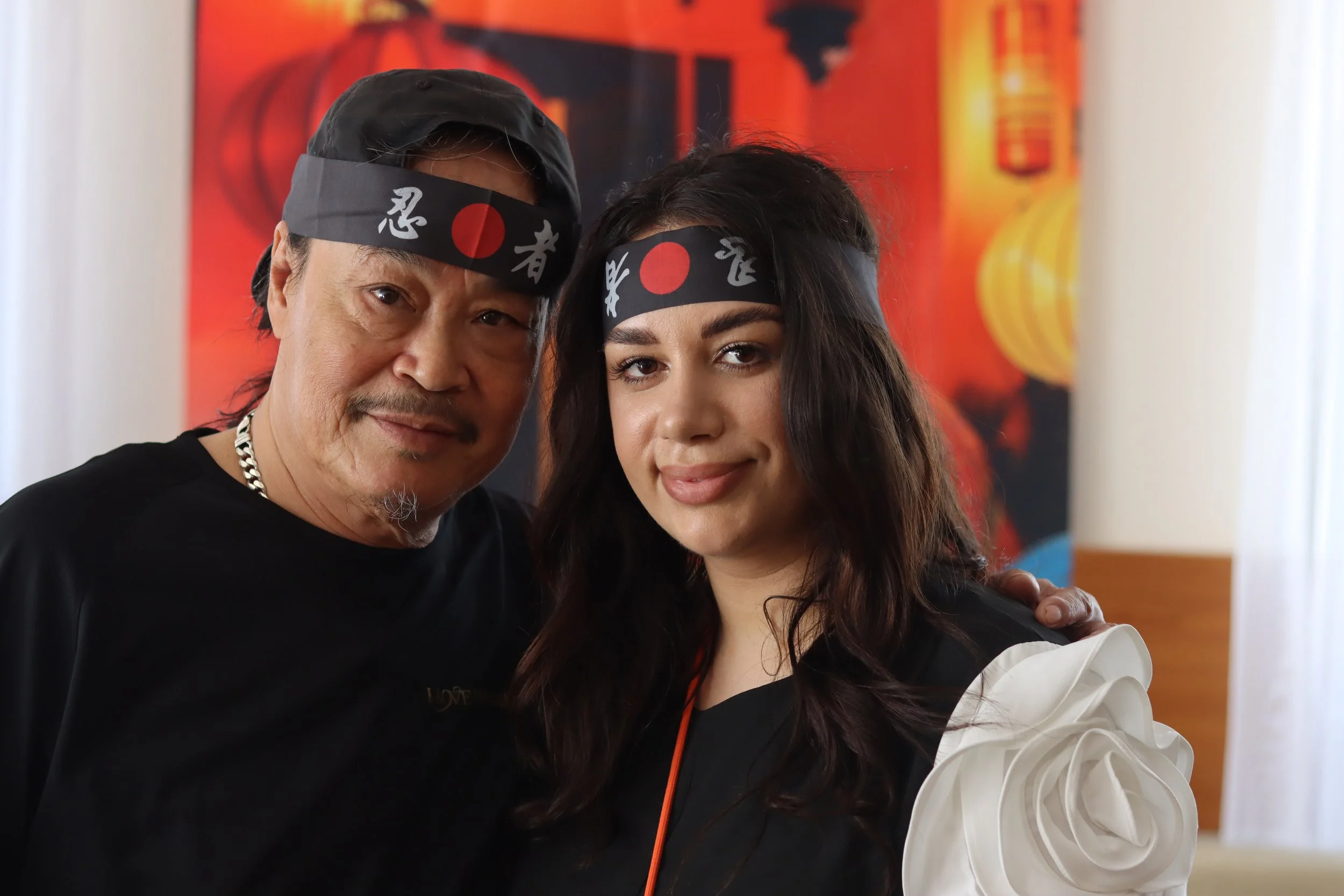 Zwei Menschen in Bandanas mit japanischen Schriftzeichen, die einander umarmen und lächeln, vor einem rot-schwarzen Hintergrund.