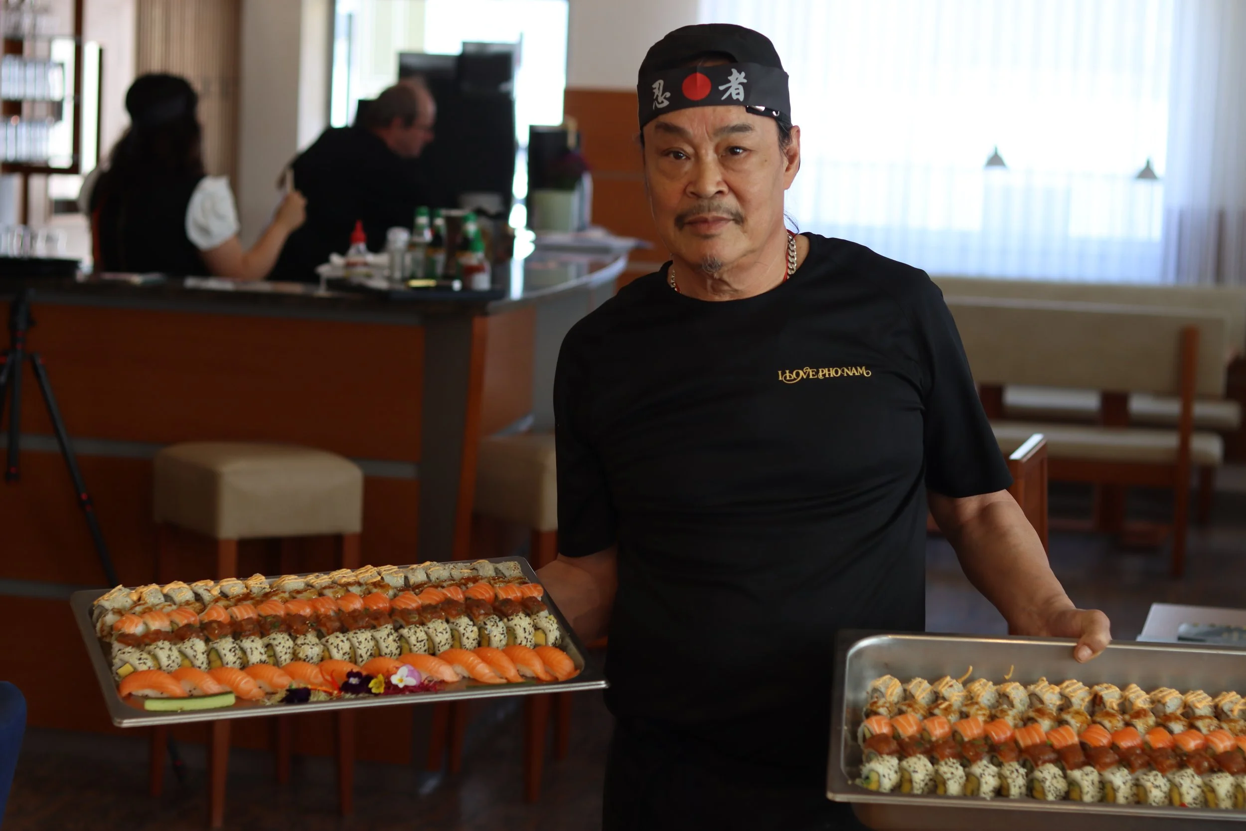 Ein Mann in schwarzer Kleidung und Hut serviert Sushi auf einem Tablett in einem Restaurant. Im Hintergrund sind zwei weitere Personen, die am Tisch essen, und große Fenster mit Vorhängen.