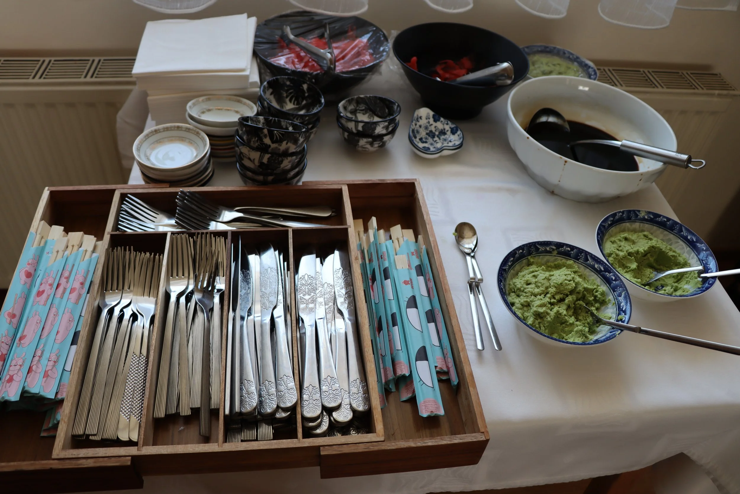 Mesa mit Sushi-Besteck, kleinen Tellern, Schüsseln mit Wasabi und Ingwer, Sojasauce, und Sushi-Zutaten.