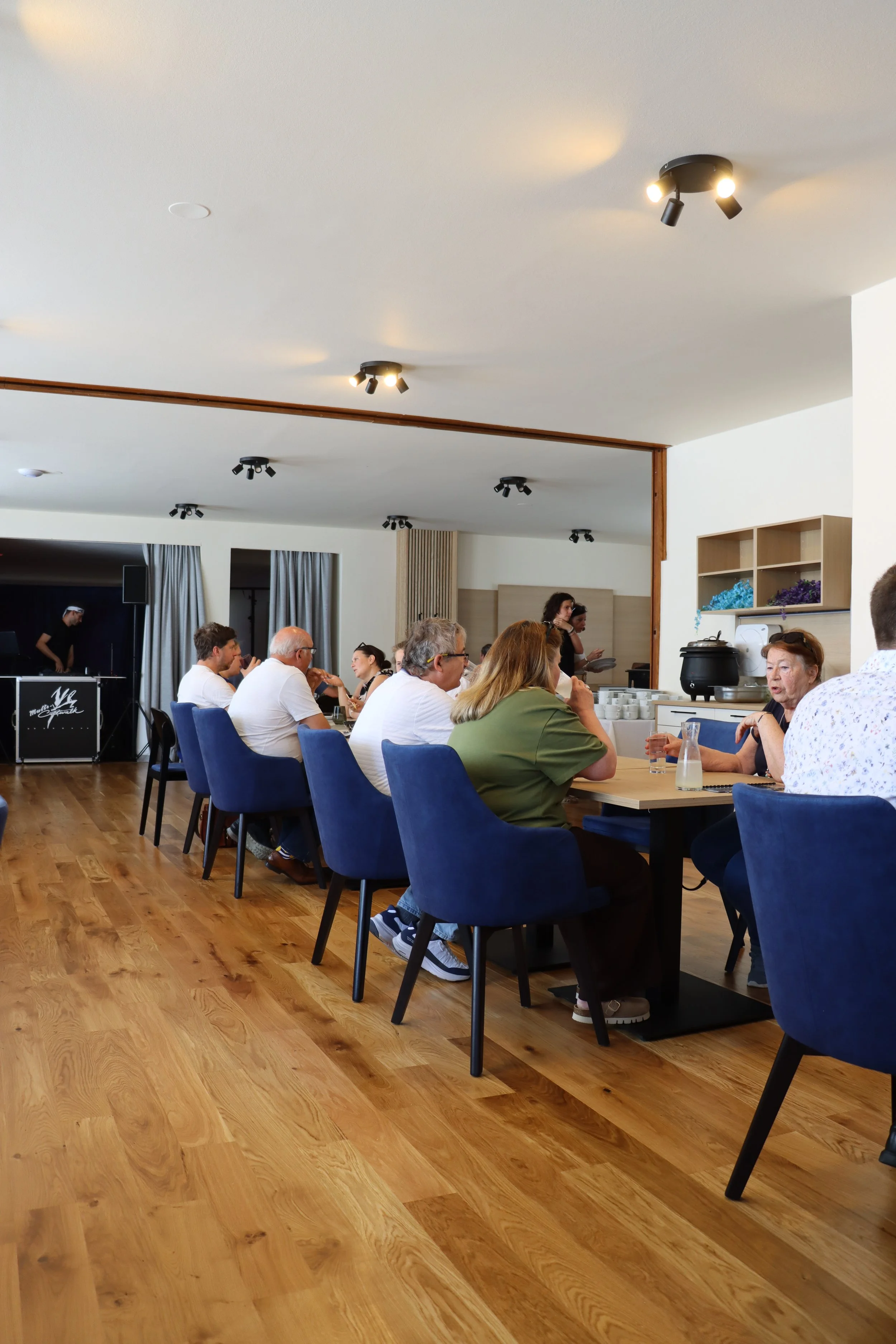 Menschen sitzen in einer modernen, gut beleuchteten Cafeteria oder Restaurant und unterhalten sich bei Getränken. Es gibt Holzfußboden, blaue Stühle und eine offene Küchen- oder Buffet-Station im Hintergrund.