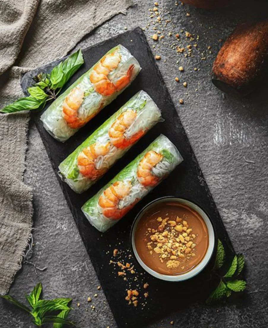 Drei vietnamesische Reisrollen mit Garnelen auf einem schwarzen Brett, daneben eine Schüssel mit brauner Sauce und zerbröseltem Erdnuss-Topping, umgeben von frischen Minzblättern.