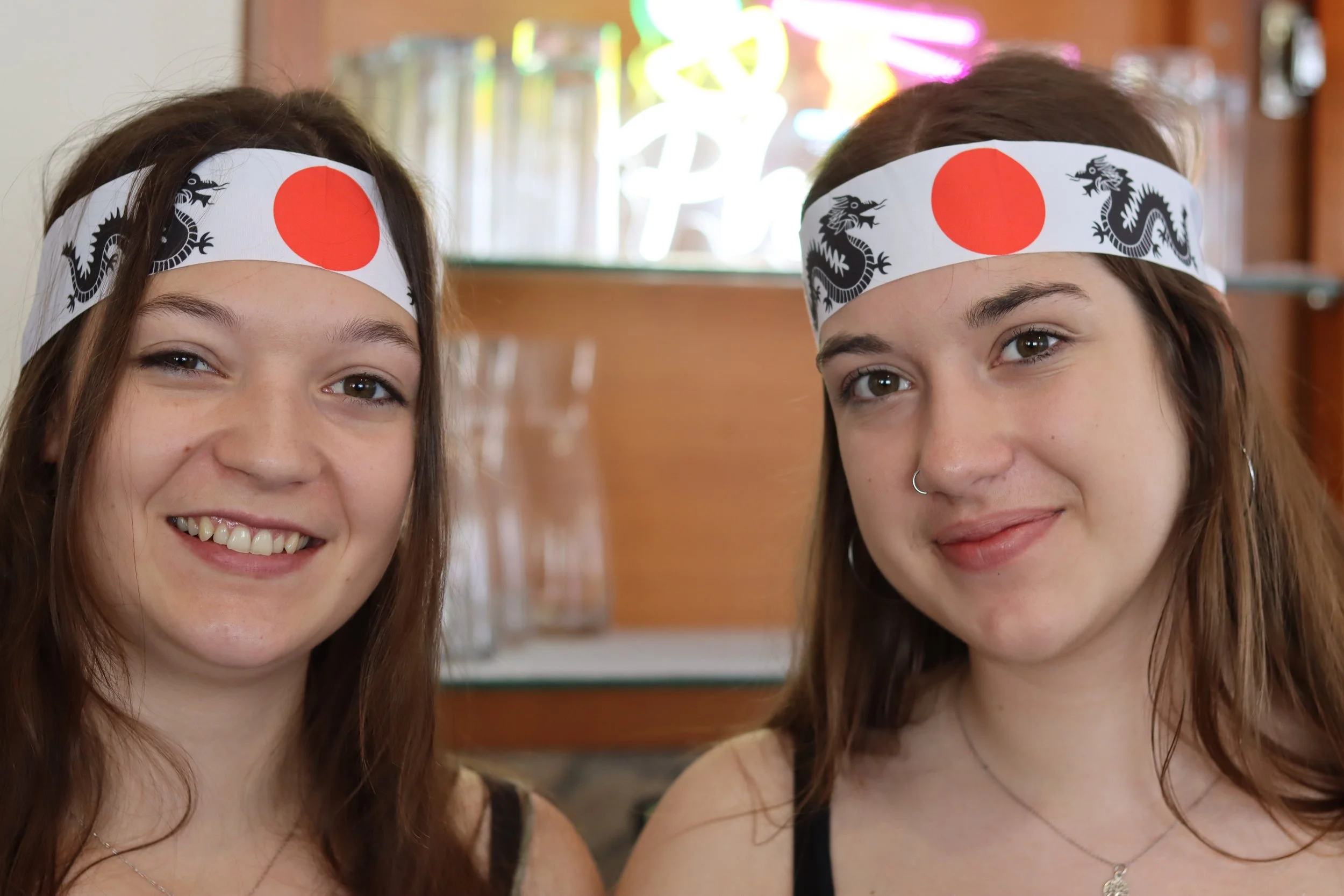 Zwei junge Frauen tragen Kopfbänder mit japanischer Flagge und Drachenmuster, lächeln, in einem Raum mit bunten Neonlichtern im Hintergrund.