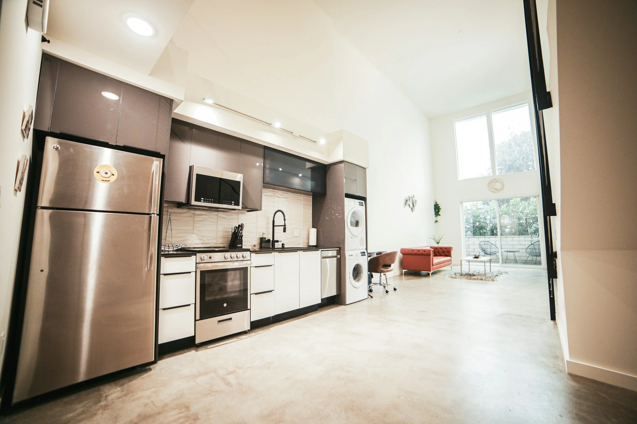 Open-concept kitchen with stainless steel appliances, white and gray cabinets, black countertop, and a small sitting area with a red couch and large window courtyard.