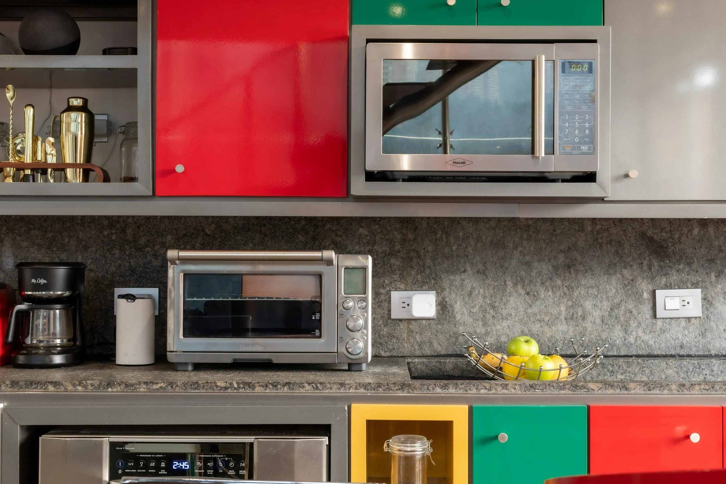 Kitchen countertop with toaster oven, coffee maker, white mug, and bowl of apples and bananas, red, green, and yellow cabinets, and various appliances.