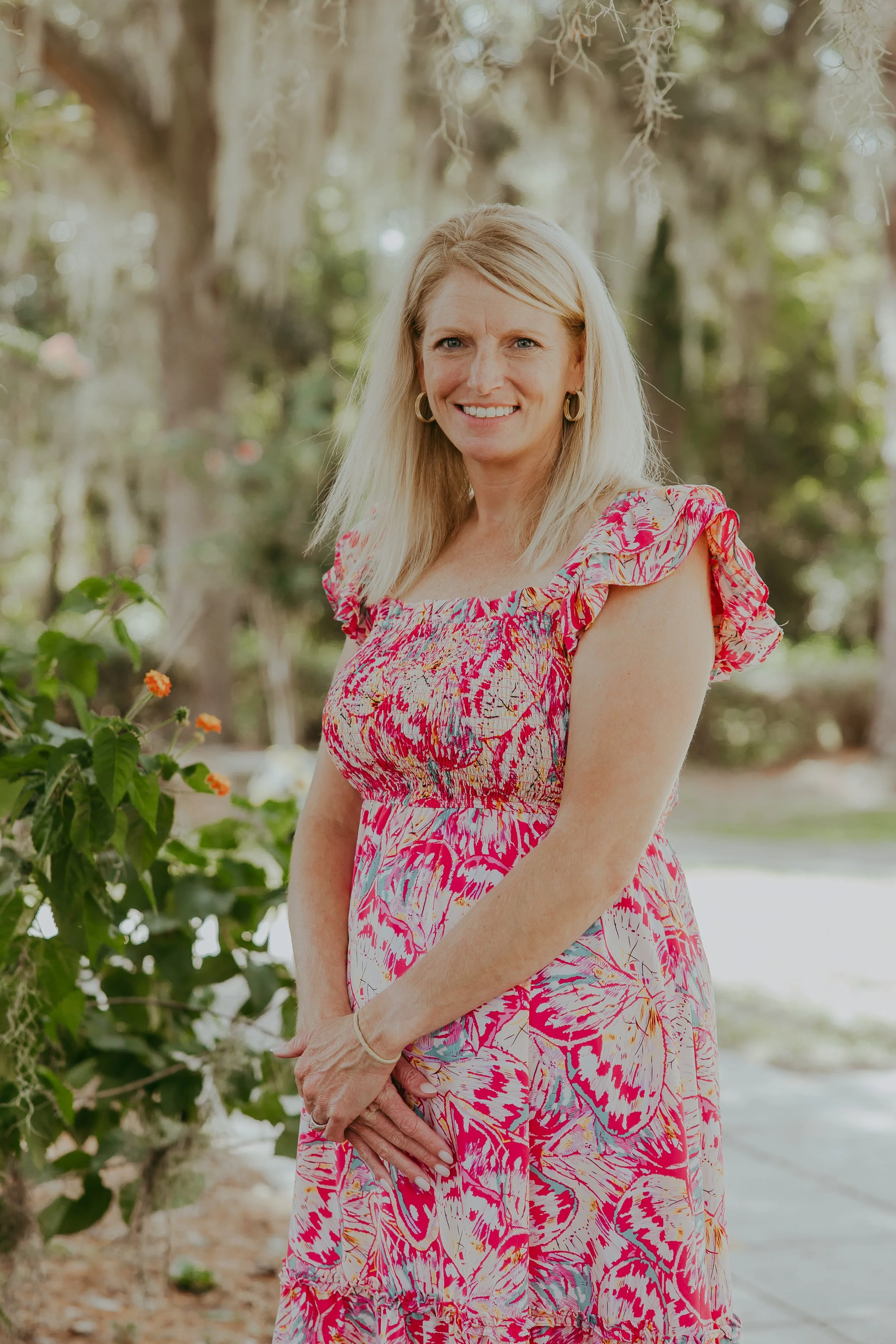 A woman with blonde hair in a floral dress and a white vest standing outdoors with trees and greenery in the background.