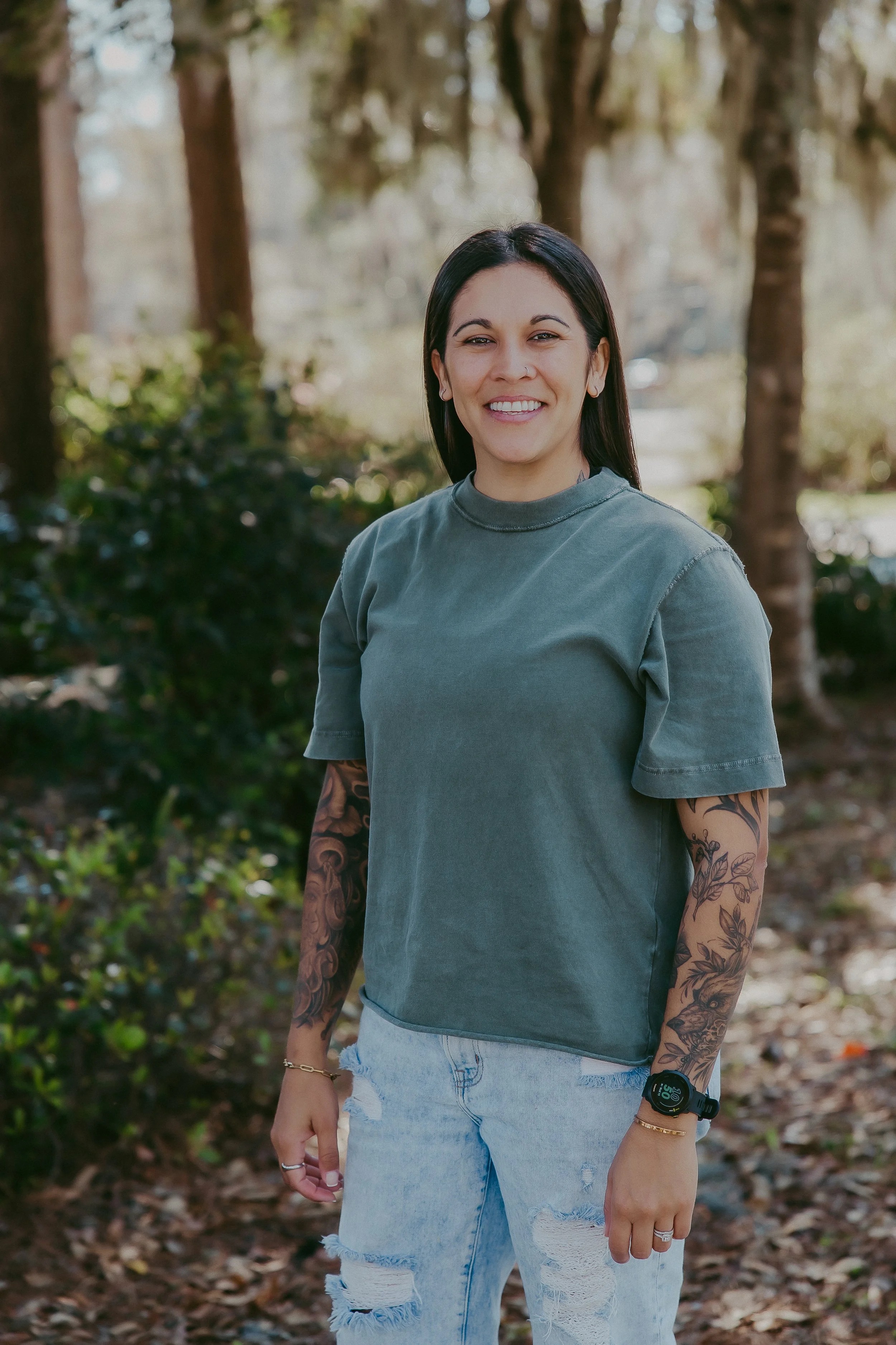 Smiling woman with dark hair, tattoos on arms, wearing a green t-shirt and ripped jeans, standing outdoors in a wooded area.