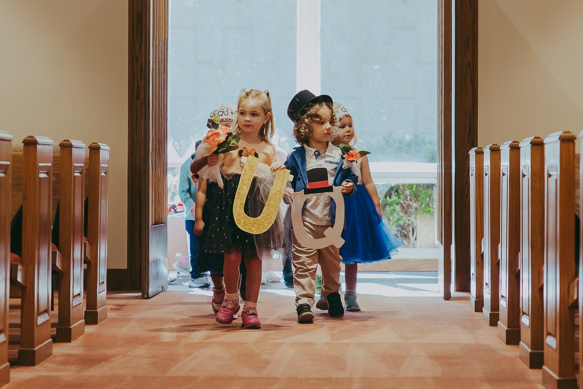 Children dressed in colorful clothes walking through an indoor hallway, holding decorated letters and flowers, with a wooden door and large window in the background.