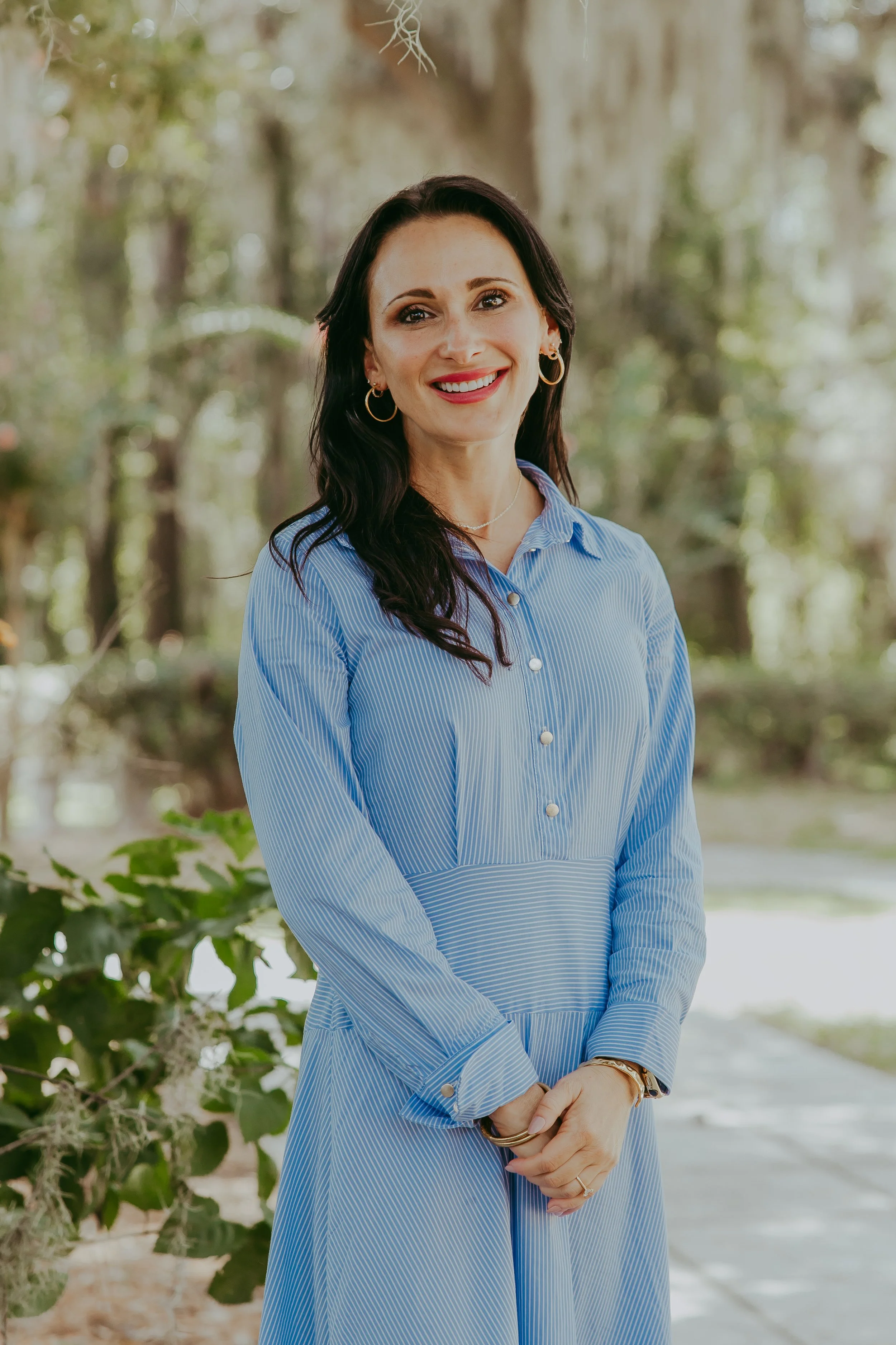 A woman with long dark hair, smiling, wearing a blue embroidered dress, standing outdoors in a lush green forest with trees and bushes.