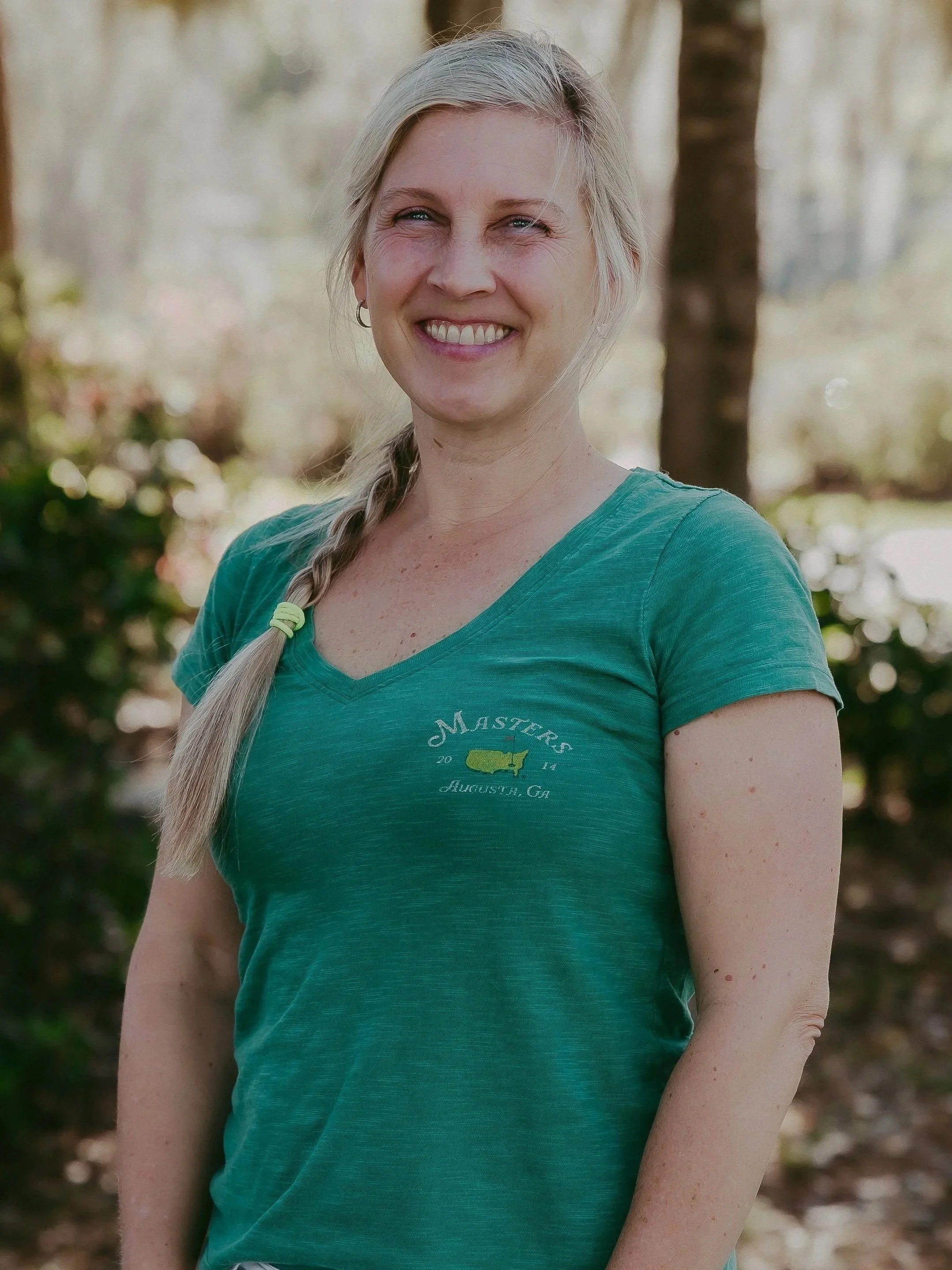 A smiling woman with blonde hair in a braid, wearing a teal t-shirt, standing outdoors with trees and blurred foliage in the background.