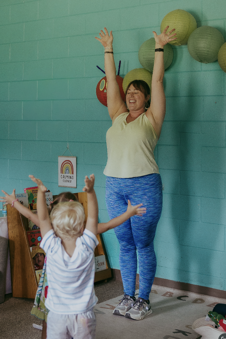 A woman in athletic wear with arms raised, smiling, in a classroom with children who are reaching out towards her, colorful paper lanterns on the wall, and a sign that says 'Calming Corner'.