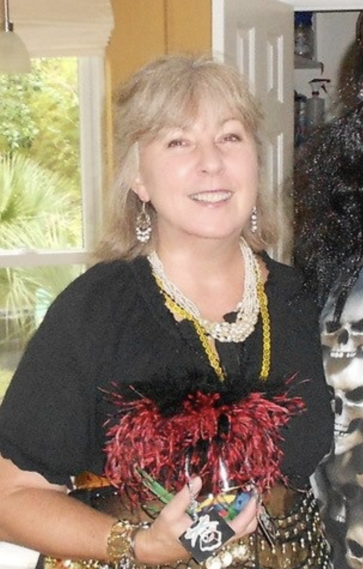 A smiling woman with shoulder-length blonde hair and earrings, wearing a black top and multi-layered necklaces, holding a red and black decorative object, standing inside a room with a window and greenery outside.