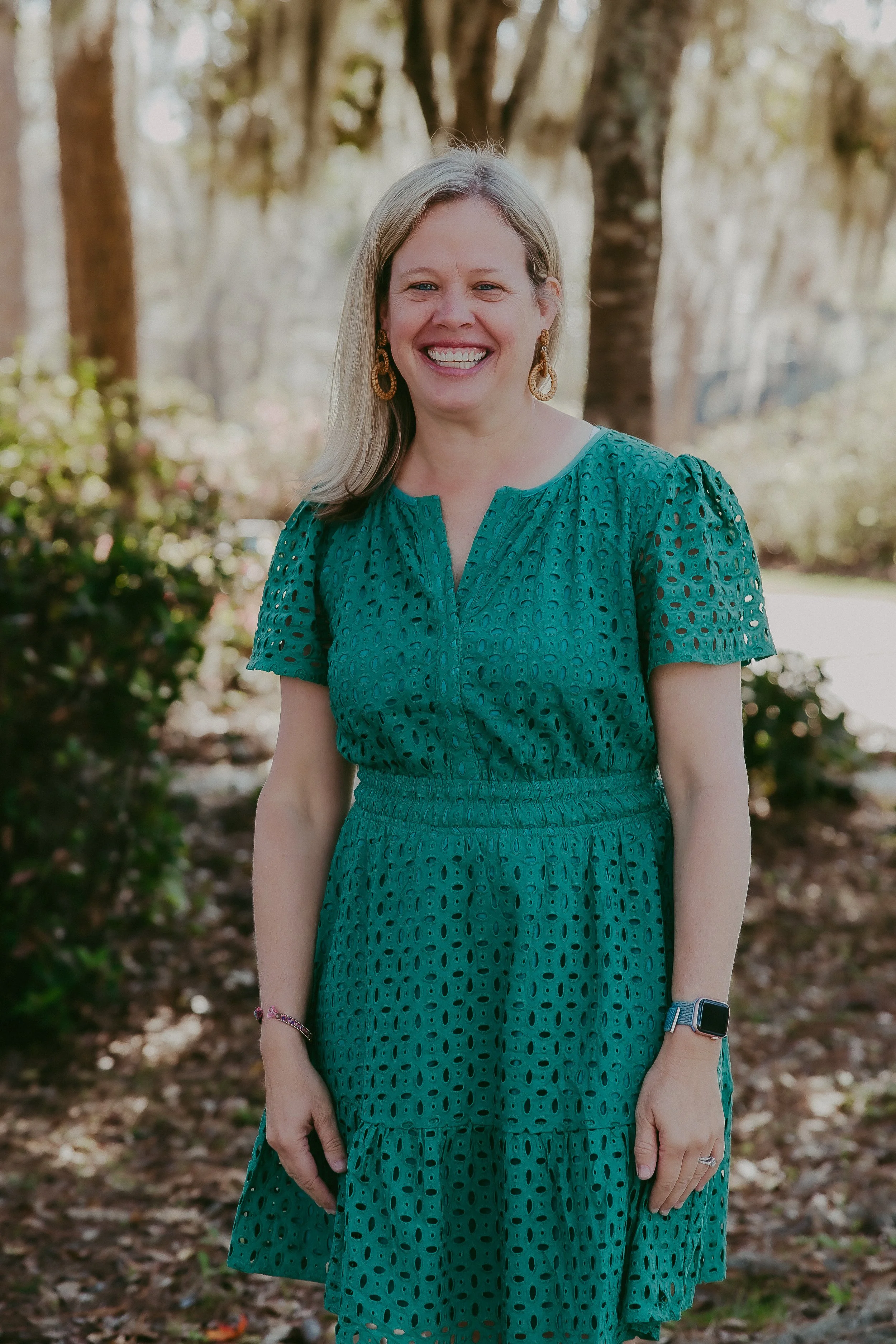 A woman smiling outdoors, wearing a teal eyelet dress and earrings, standing among trees and bushes.