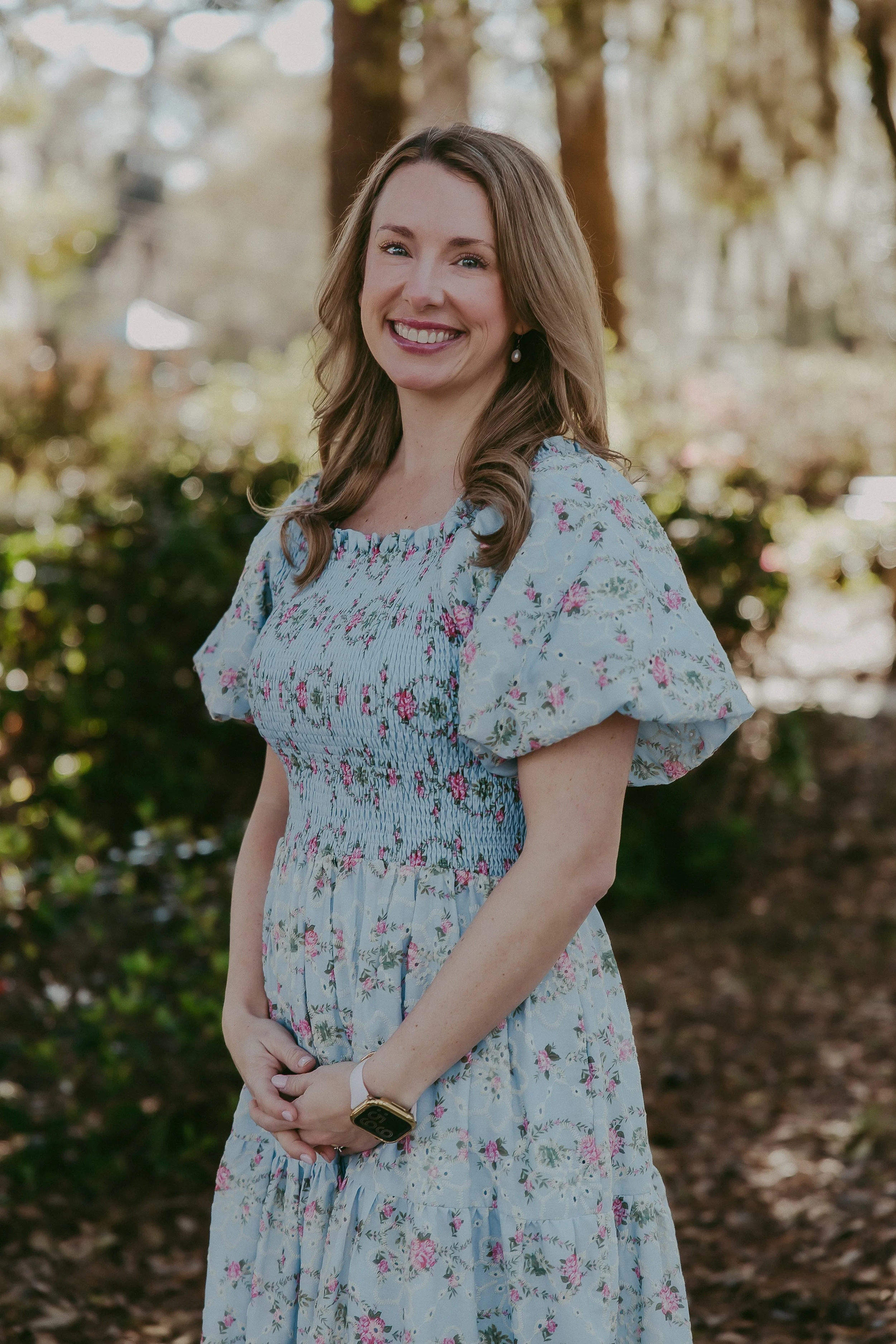 A woman with wavy brown hair, wearing a light blue floral dress with puffed sleeves, standing outdoors and smiling at the camera.
