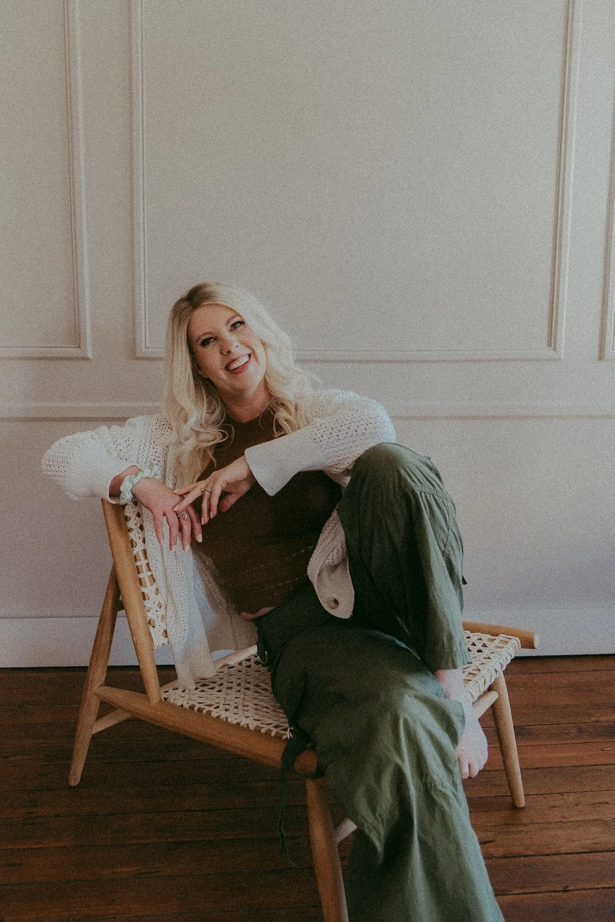 A woman with long blonde wavy hair wearing a beige beanie, a tan top, and a red plaid shirt holding a to-go coffee cup at the beach, laughing cheerfully.