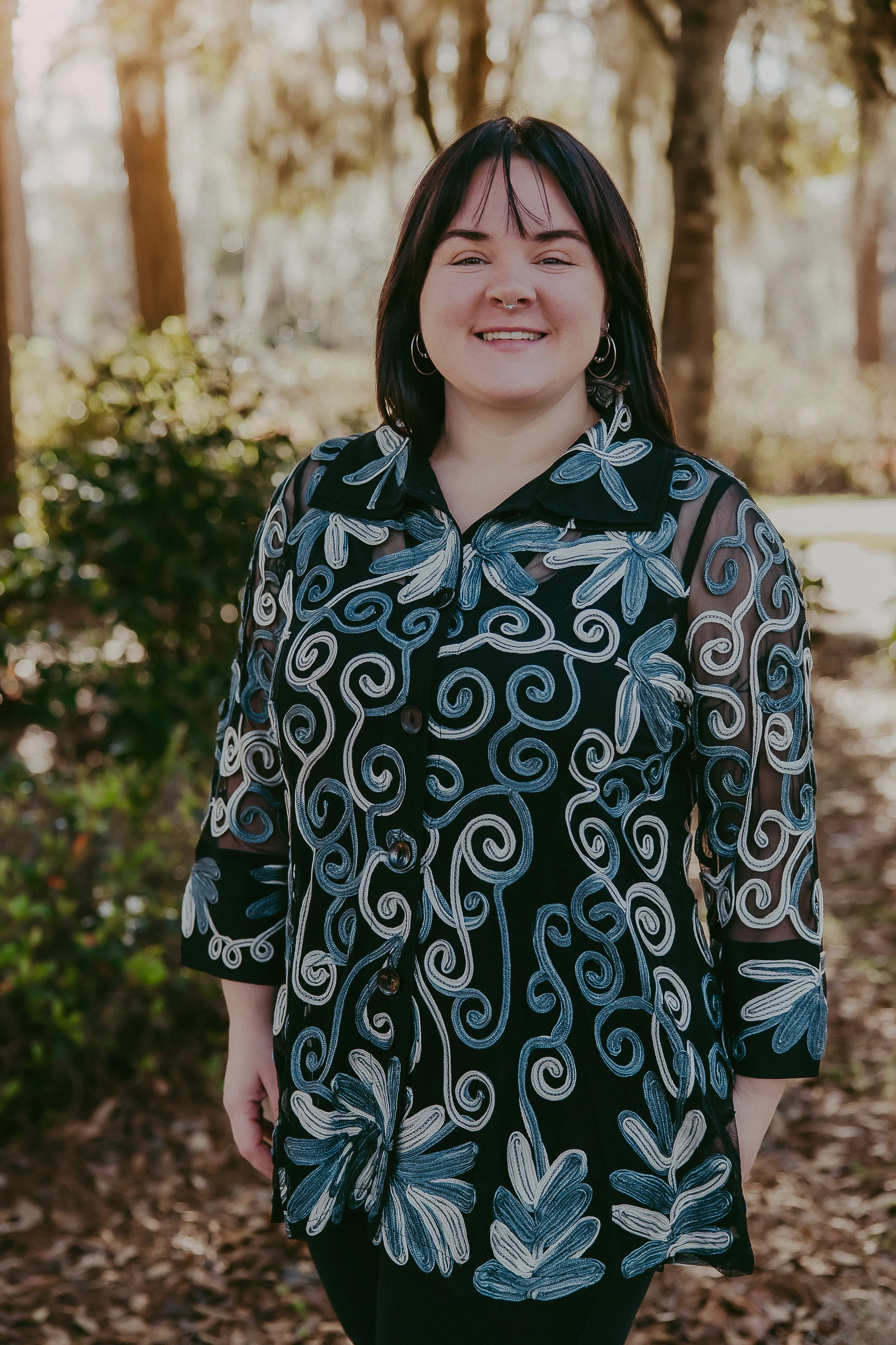 A smiling woman with black hair, wearing a sheer black blouse with blue and white embroidered floral and swirl patterns, standing outdoors in a wooded area.