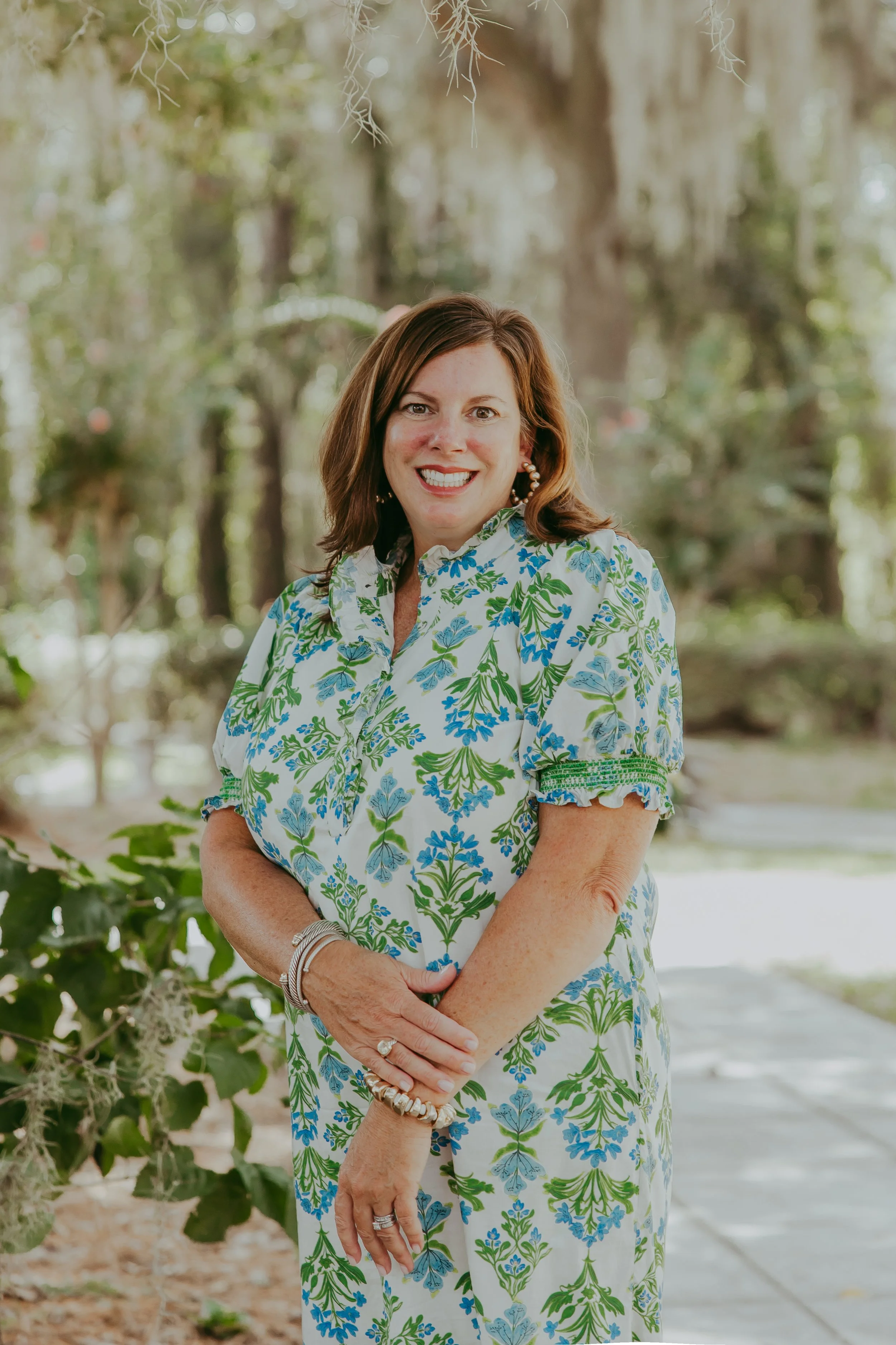 A woman smiling outdoors in front of trees and greenery, wearing a white coat and gold jewelry.