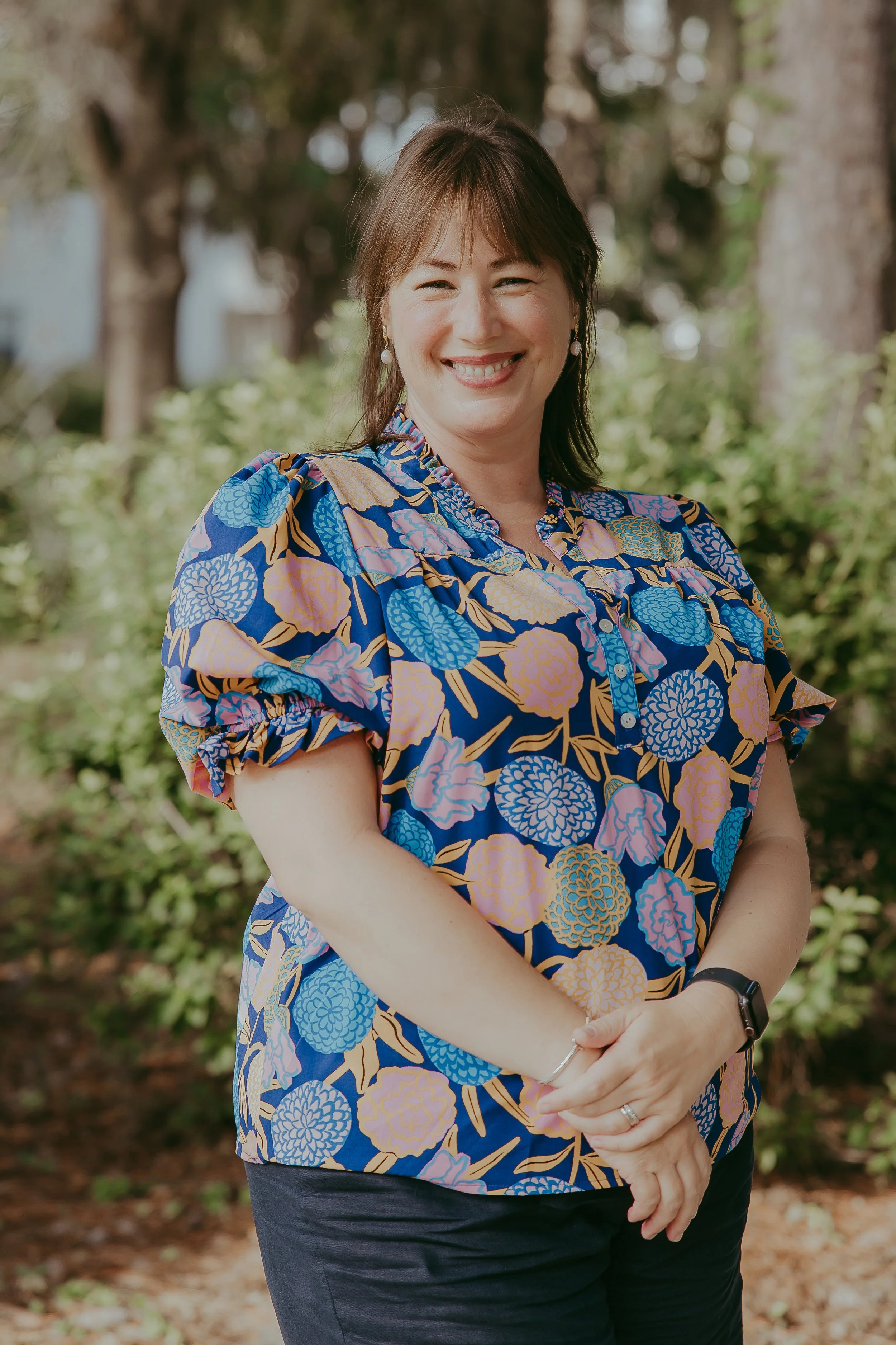 A woman smiling outdoors, wearing a colorful floral blouse with blue, pink, and yellow flowers, and dark pants.